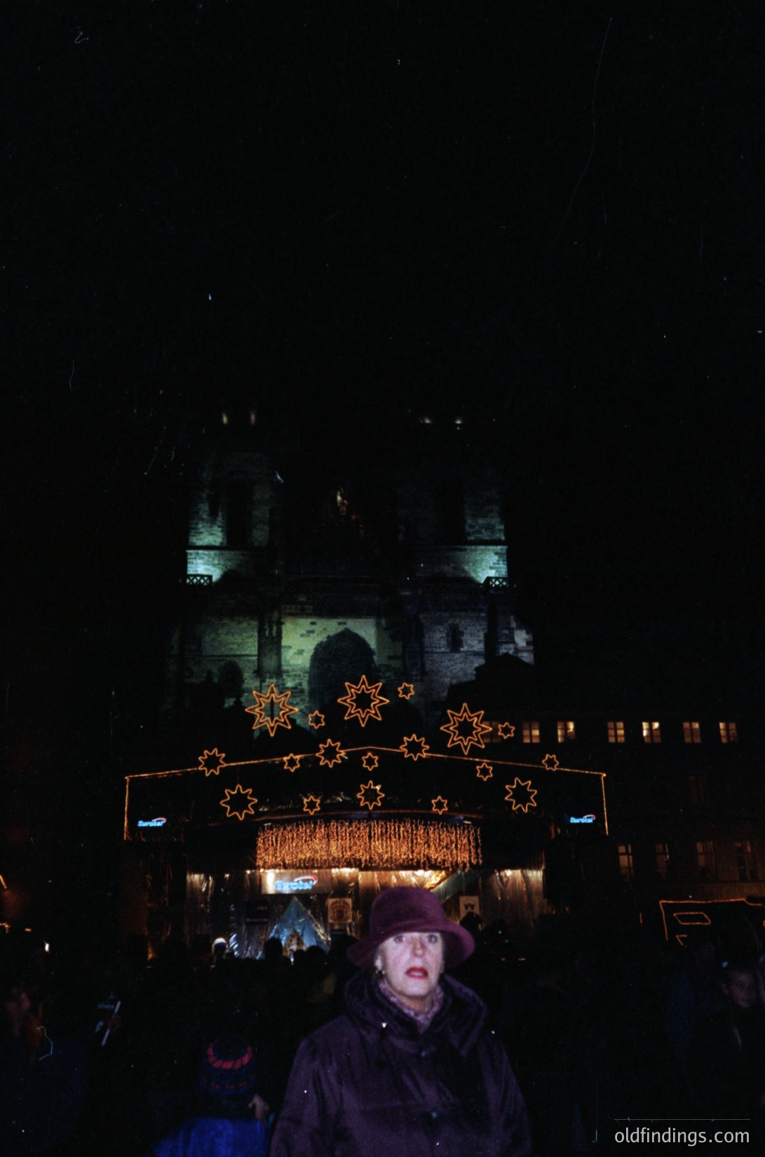 Nighttime winter festival scene featuring illuminated star decorations and a historic stone building. Warm golden lights contrast with cold shadows, highlighting architectural details. Crowd in winter attire suggests chilly weather.