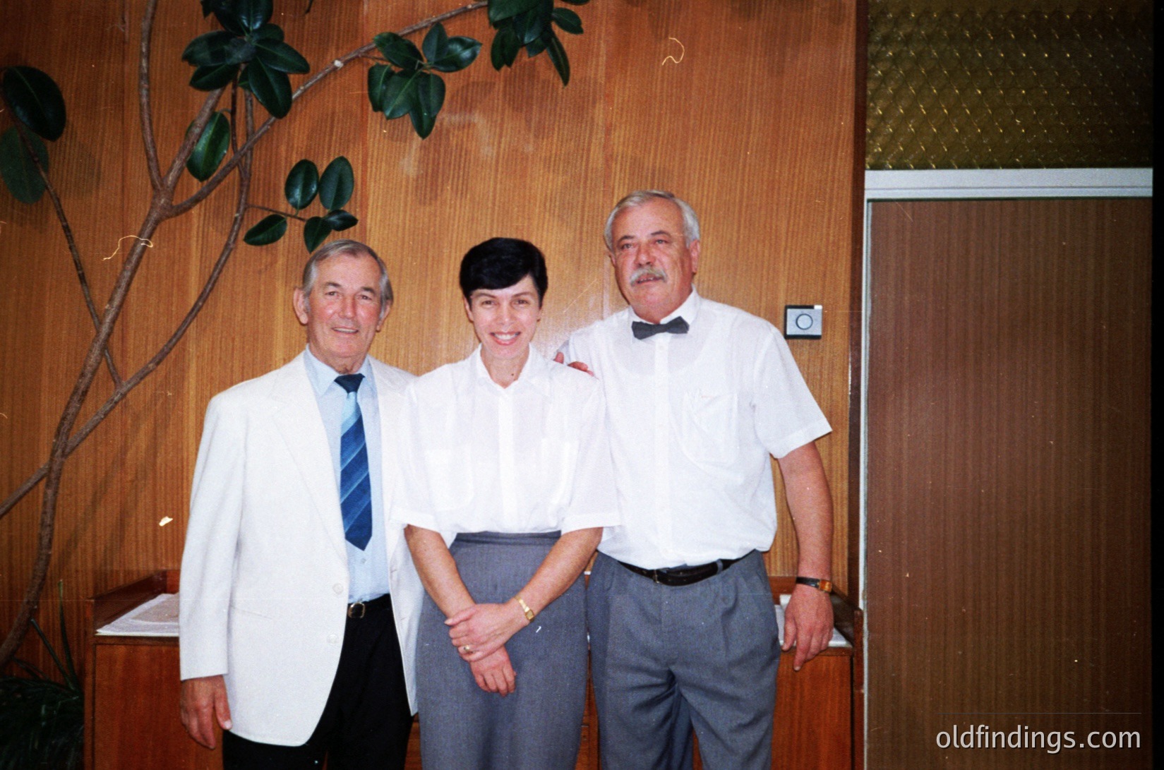 Three adults pose indoors in formal attire, likely mid-1990s. Left: man in white blazer, striped tie. Center: woman in light blouse, dark skirt. Right: man in white shirt, bow tie, gray trousers. Wood-paneled walls, potted plant, and door with metal handle visible. Warm, professional setting.
