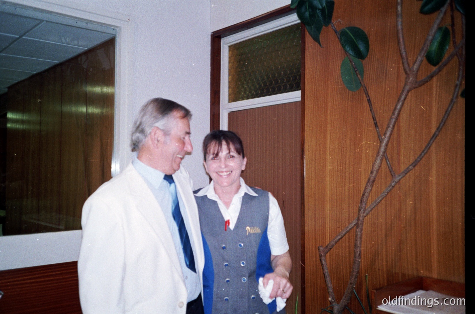 Two individuals in a formal indoor setting, likely a 1980s-1990s office or hotel lobby. The man wears a white lab coat and striped tie; the woman, a blue-and-white "Pension" uniform with a red badge. Wood-paneled walls and a potted plant frame the scene.