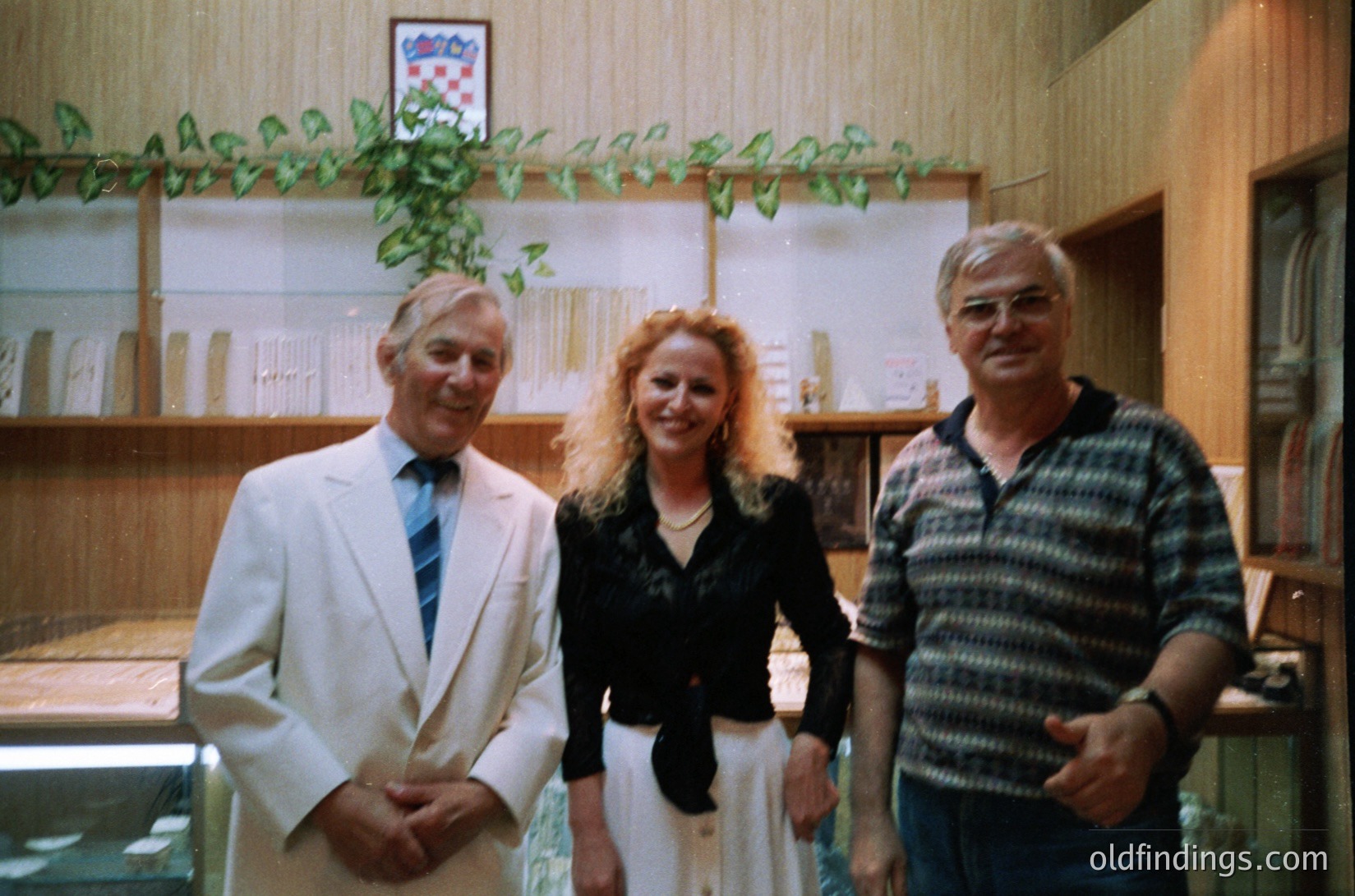 Three individuals pose indoors in a formal setting, likely mid-1970s–1980s. The man on the left wears a white suit with a striped tie, the woman a dark blazer over a light dress, and the man on the right a patterned polo shirt. Wooden shelving with framed items and a potted plant with green leaves frame the background. Warm lighting and vintage color tones suggest a professional or cultural event.