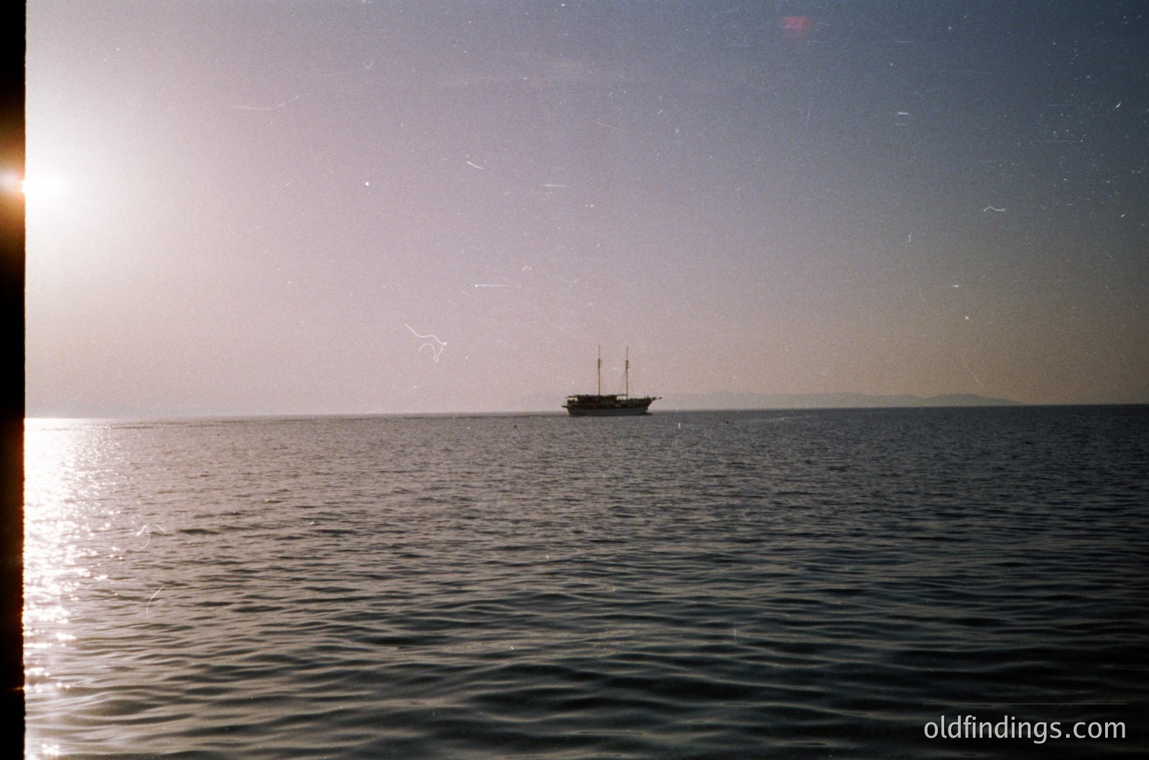 Vintage seascape shot of a single-masted wooden sailing vessel on calm waters, framed by a window sill. Soft, grainy film grain suggests mid-20th century (1950s–1970s). Distant shoreline and hazy horizon imply coastal Mediterranean or Atlantic setting.