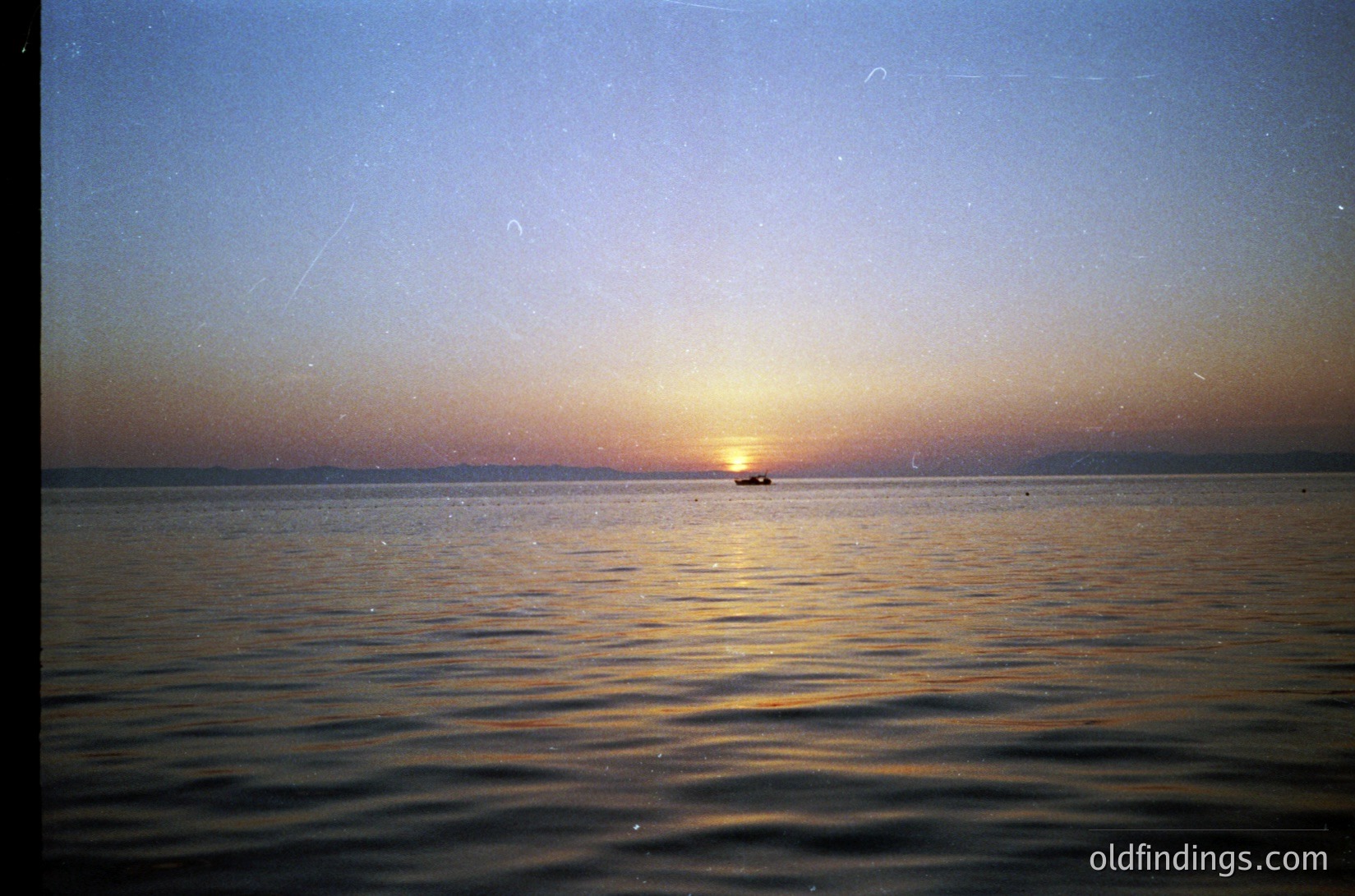 Golden sunset over calm waters with a lone boat silhouetted near horizon. Warm gradient sky transitions from deep blue to orange. Likely captured with vintage film—soft focus and slight grain. Evokes serene coastal or lake setting.