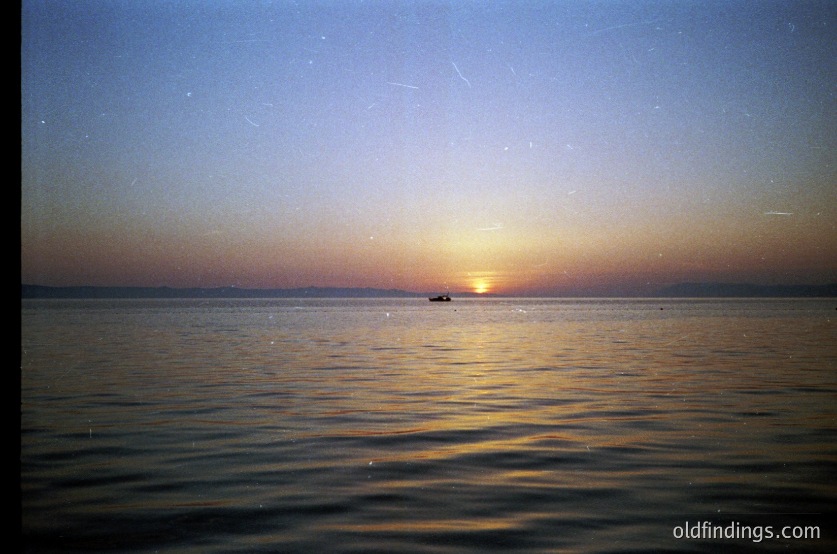 Sunset over calm waters with a lone boat silhouetted near horizon. Warm golden light reflects on rippling sea, framed by fading twilight sky. Vintage film grain suggests mid-20th century photography. Ideal for mood-driven stock or travel inspiration.