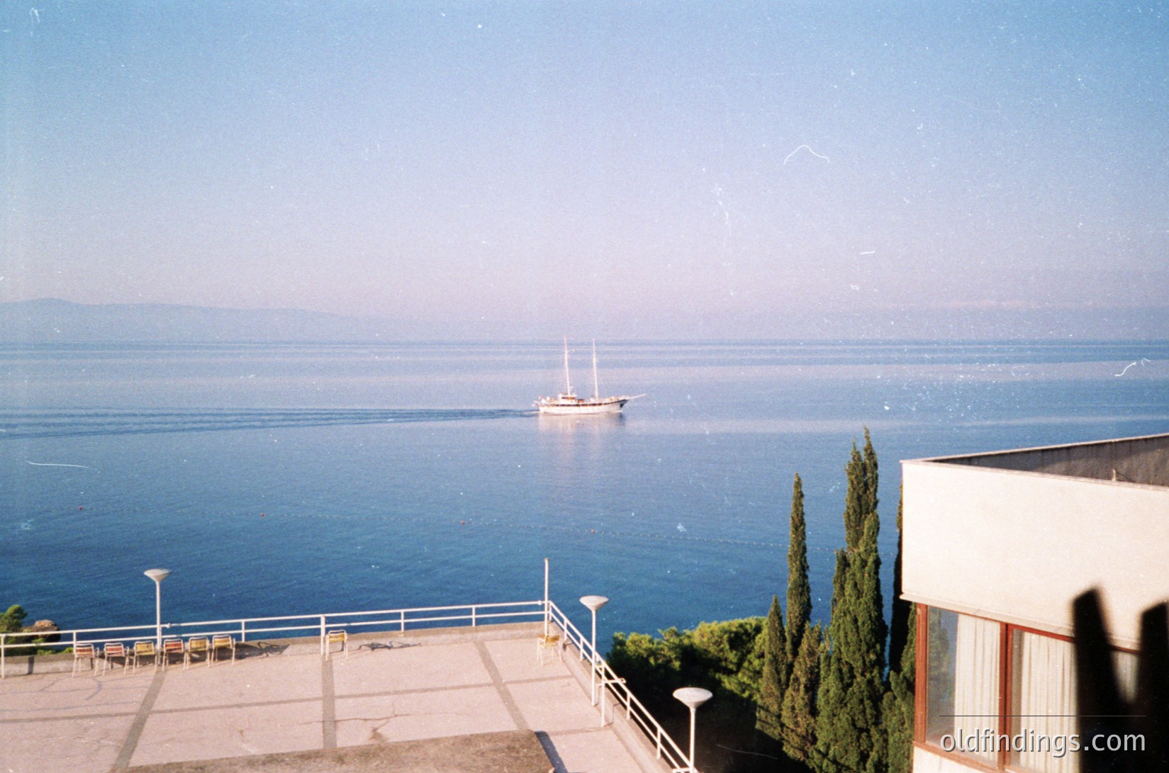 Vintage seaside view of calm waters with a single-masted sailboat anchored. Concrete terrace with curved railings and potted cypress trees frames the composition. Mid-20th century coastal architecture visible at right. Soft vintage filter suggests 1960s–1980s era.