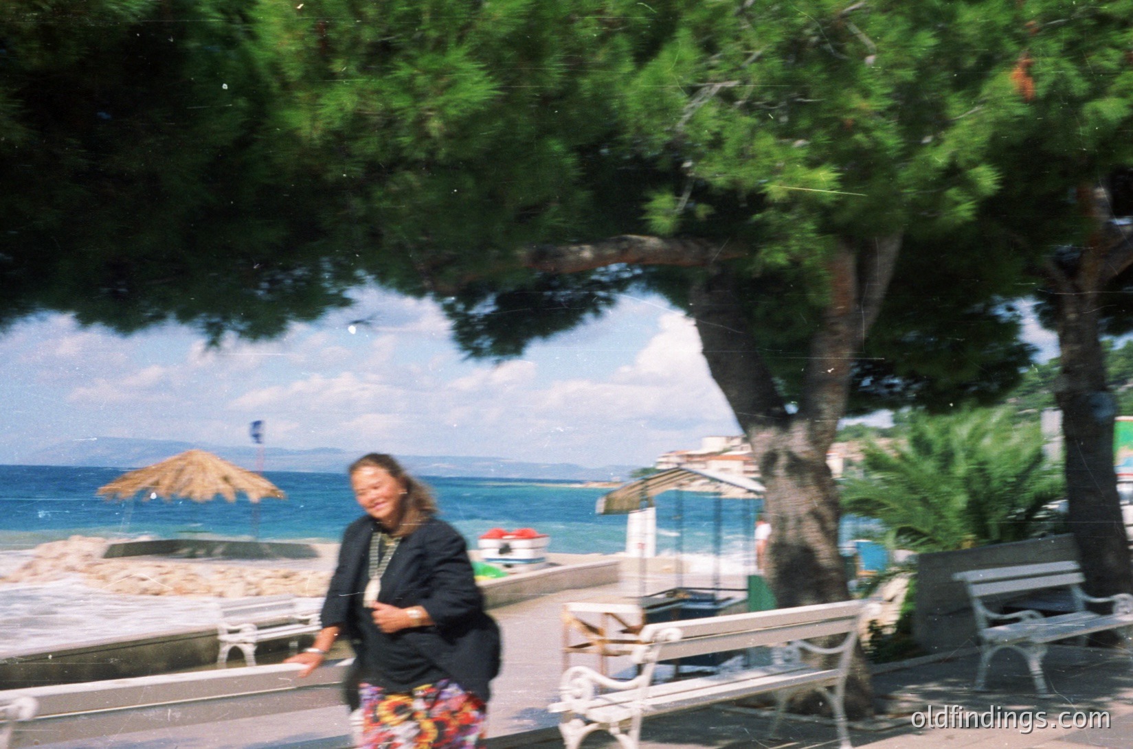 Vintage seaside scene featuring a woman in a patterned dress walking under a large tree, with ocean and beach umbrellas in background. Likely Mediterranean resort, 1970s–1990s.