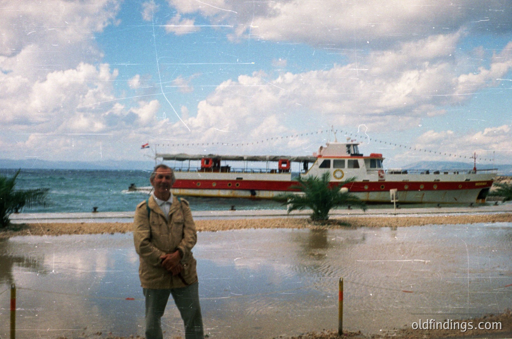 Vintage seaside scene featuring a man in a beige trench coat by a shallow, reflective waterfront. A red-and-white ferryboat with decorative string lights and a flag adorns the background, suggesting a festive or tourist setting. Coastal landscape with distant mountains under a partly cloudy sky. Likely mid-20th century, Eastern European seaside resort.