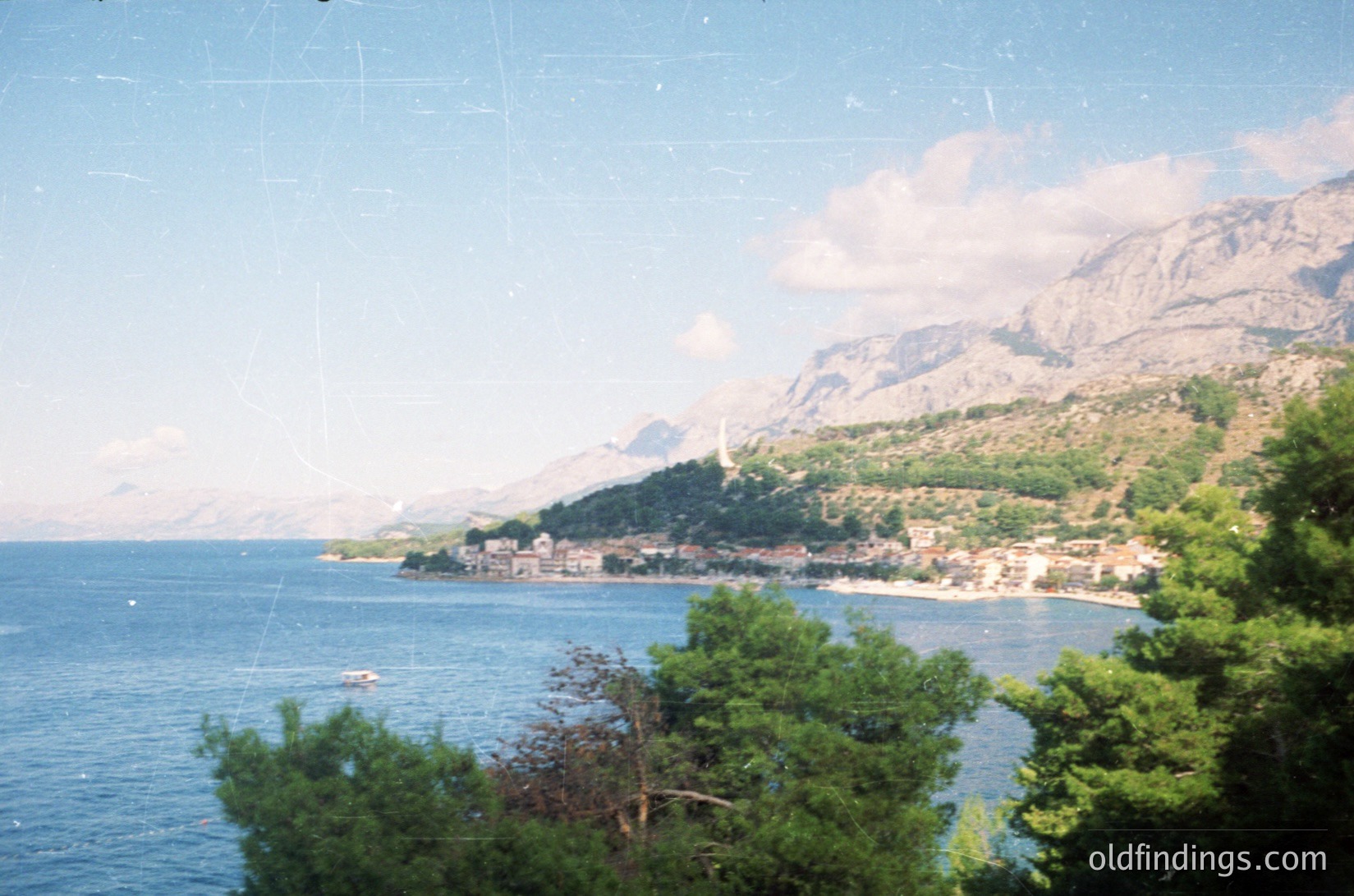 Vintage coastal scene featuring a serene bay framed by lush greenery and rugged mountains. A small coastal village with clustered buildings lines the shore, overlooking calm waters. The image captures a vintage aesthetic, likely from the **1980s-1990s**, with a soft focus and slight grain.