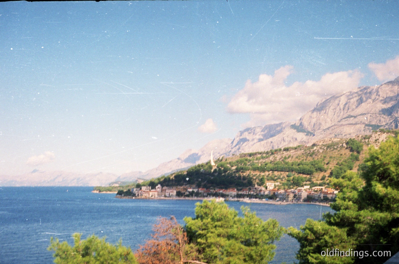 Coastal town nestled between deep blue waters and rugged mountains, likely Mediterranean. Dense greenery frames the foreground, with a church spire visible among clustered buildings. Vintage film grain suggests 1980s–1990s era. Ideal for travel inspiration or historical geography studies.