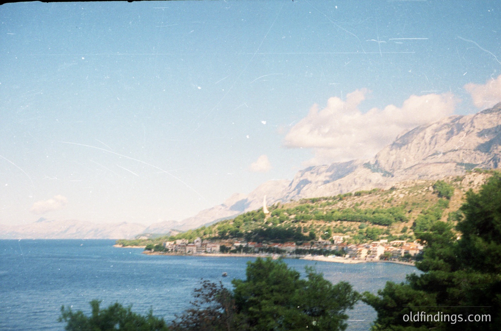 Vintage coastal scene with steep forested mountains framing a small seaside village. Dense greenery meets turquoise waters under clear skies, suggesting Mediterranean geography. Likely 1980s–1990s due to film grain and clothing style.