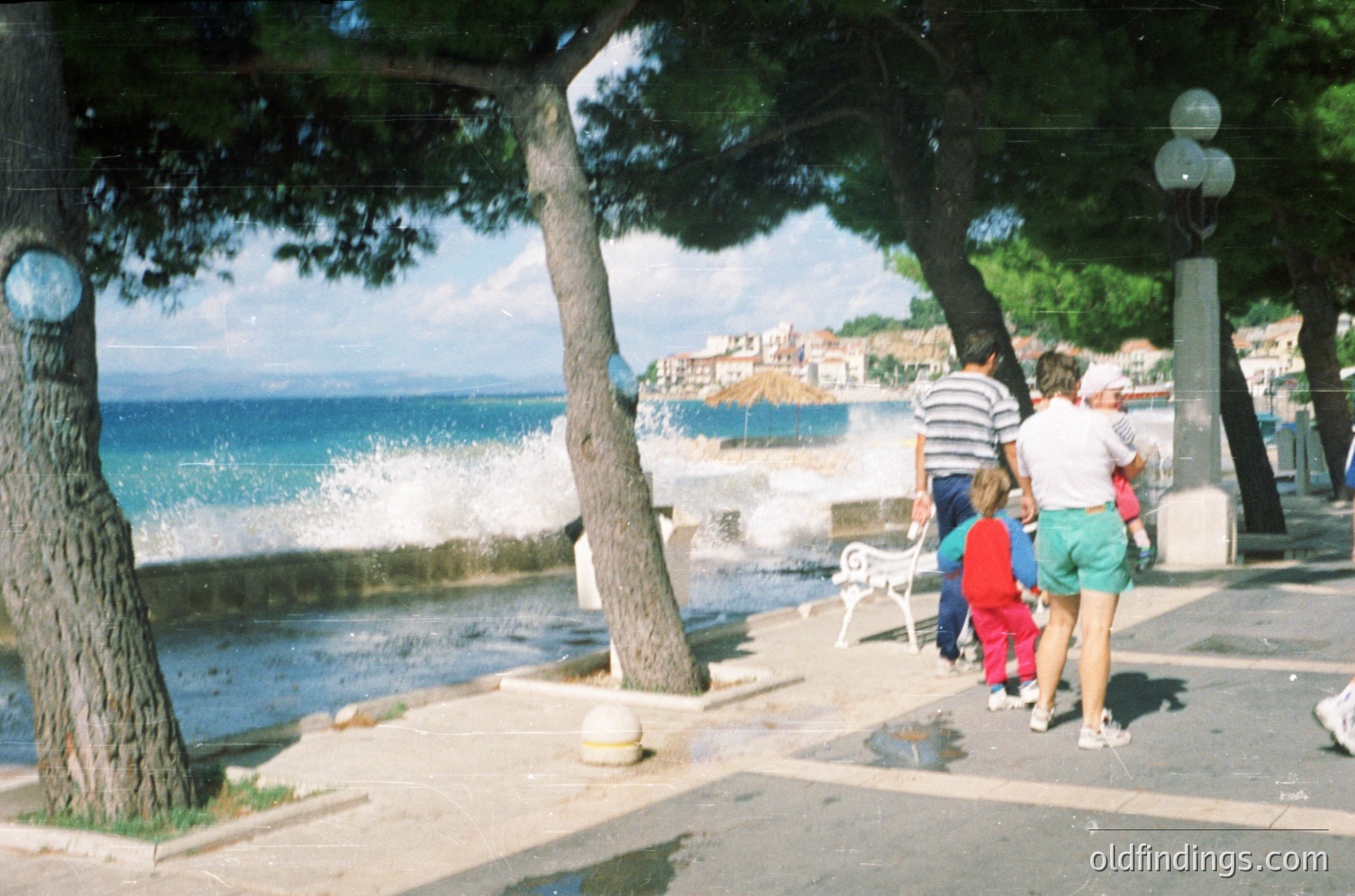 Family strolls along a seaside promenade with Mediterranean coastal town in background. Lush pine trees frame the scene, while waves crash onto rocks below. Casual summer attire suggests 1990s–2000s era.