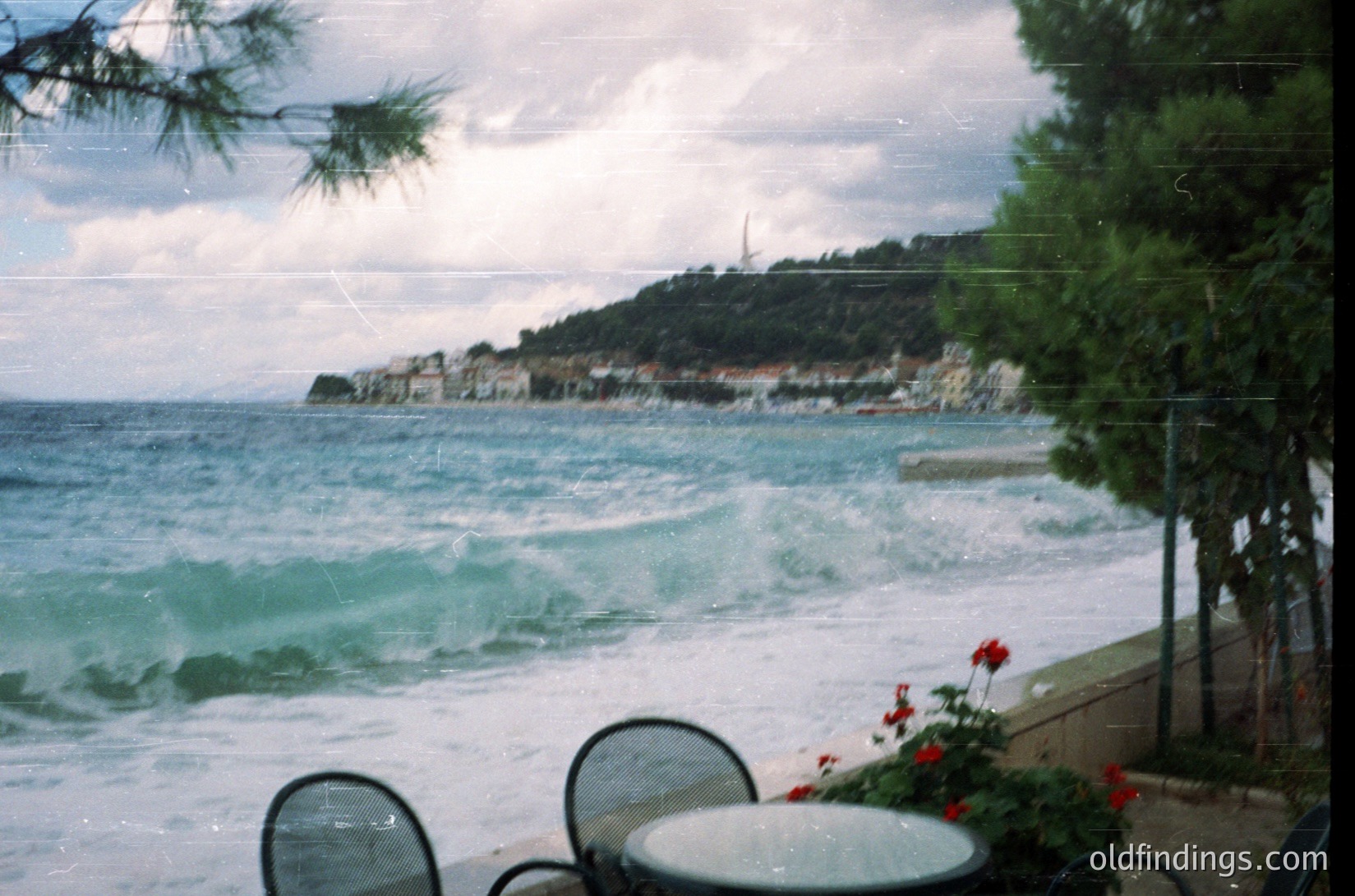 Seaside view through vintage glass patio doors, featuring crashing waves on a rocky coastline. Metal patio furniture and red flowers frame the scene, suggesting a coastal café or resort. Overcast skies enhance the dramatic ocean texture. Likely Mediterranean or Black Sea region, mid-20th century. é
