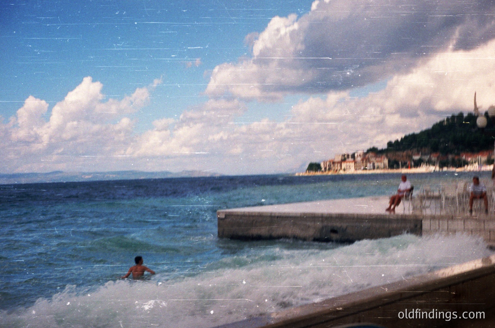 Vintage seaside scene with a man swimming in shallow waves near a concrete pier. Two figures sit on the edge, overlooking the water. Lush greenery and distant buildings line the horizon under a partly cloudy sky. Likely Mediterranean coastal area, 1960s–1980s.