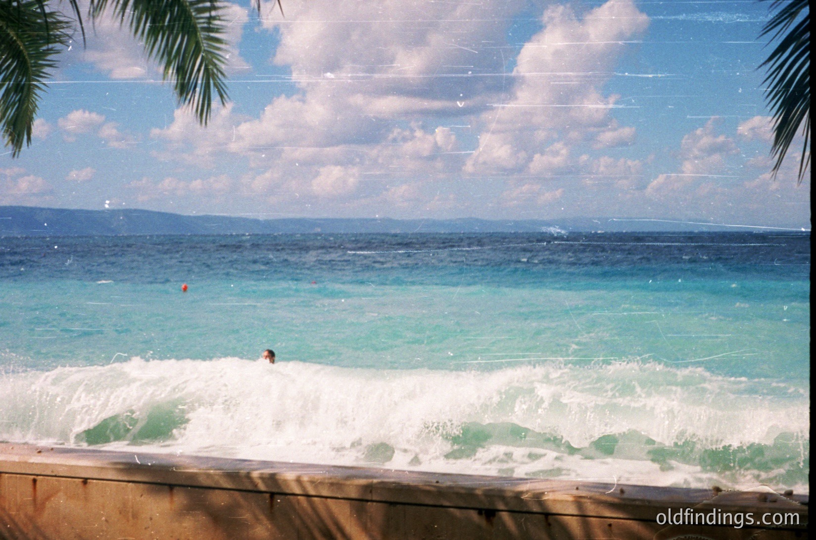 Vibrant turquoise waves crashing near a palm-fringed pool edge, with a lone surfer riding a small wave. Bright blue ocean meets fluffy clouds under clear skies. Likely a resort or coastal retreat, mid-20th century tropical setting.