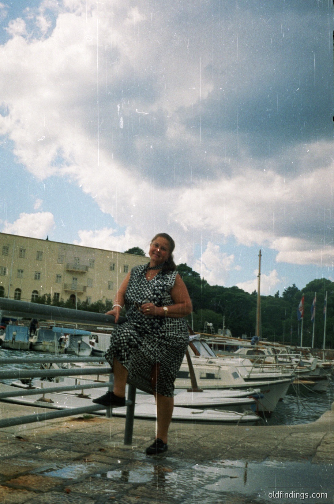 Vintage 1970s-style polka-dot dress with high-neck and puff sleeves, paired with knee-high boots. Woman poses on wet pavement beside marina with moored sailboats. Mid-rise concrete buildings and greenery in background under dramatic storm clouds.