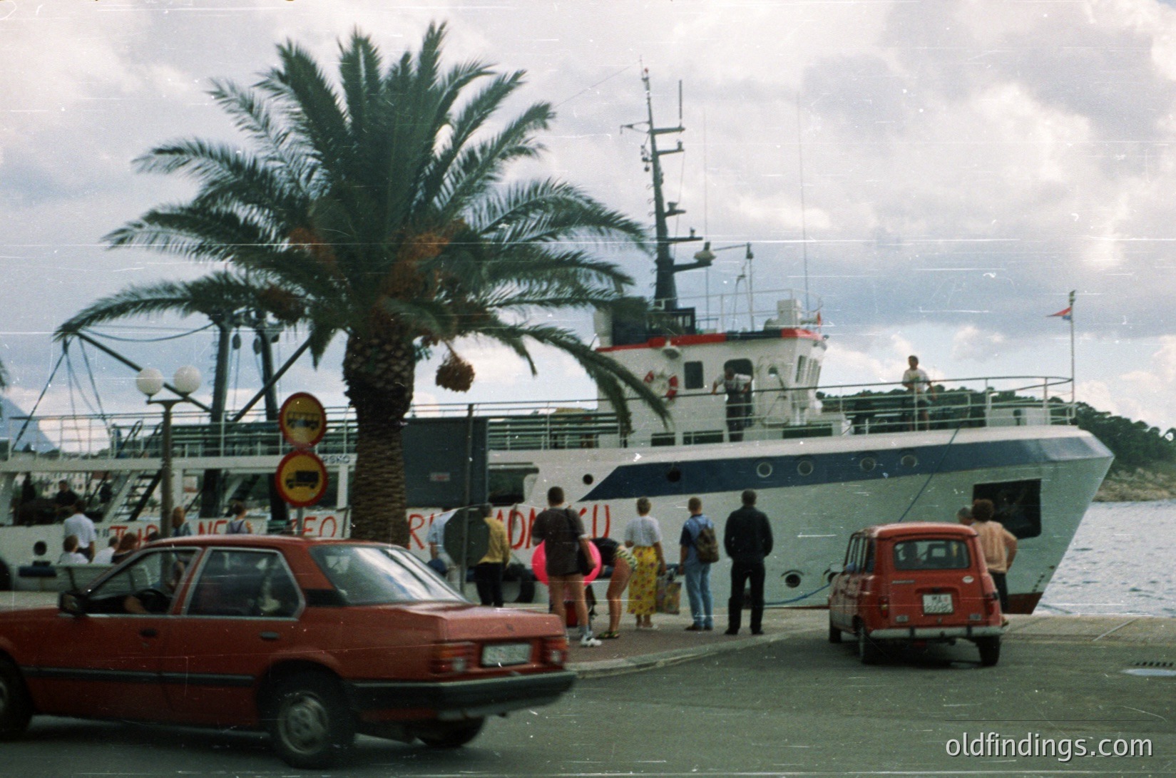 Mid-20th century ferry port scene with vintage ferry, palm tree, and classic cars. Passengers gather near boarding ramp; red cars parked nearby. Likely Mediterranean coastal area, 1960s-1970s.