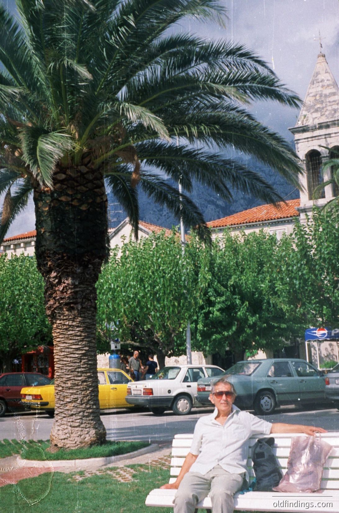 A man sits on a concrete bench beside a palm tree in a Mediterranean-style plaza, overlooking a historic building with arched windows and terracotta roof tiles. Yellow taxis and older European cars fill the street, suggesting a coastal city during the 1980s-1990s. The scene captures urban greenery, vintage vehicles, and architectural details.