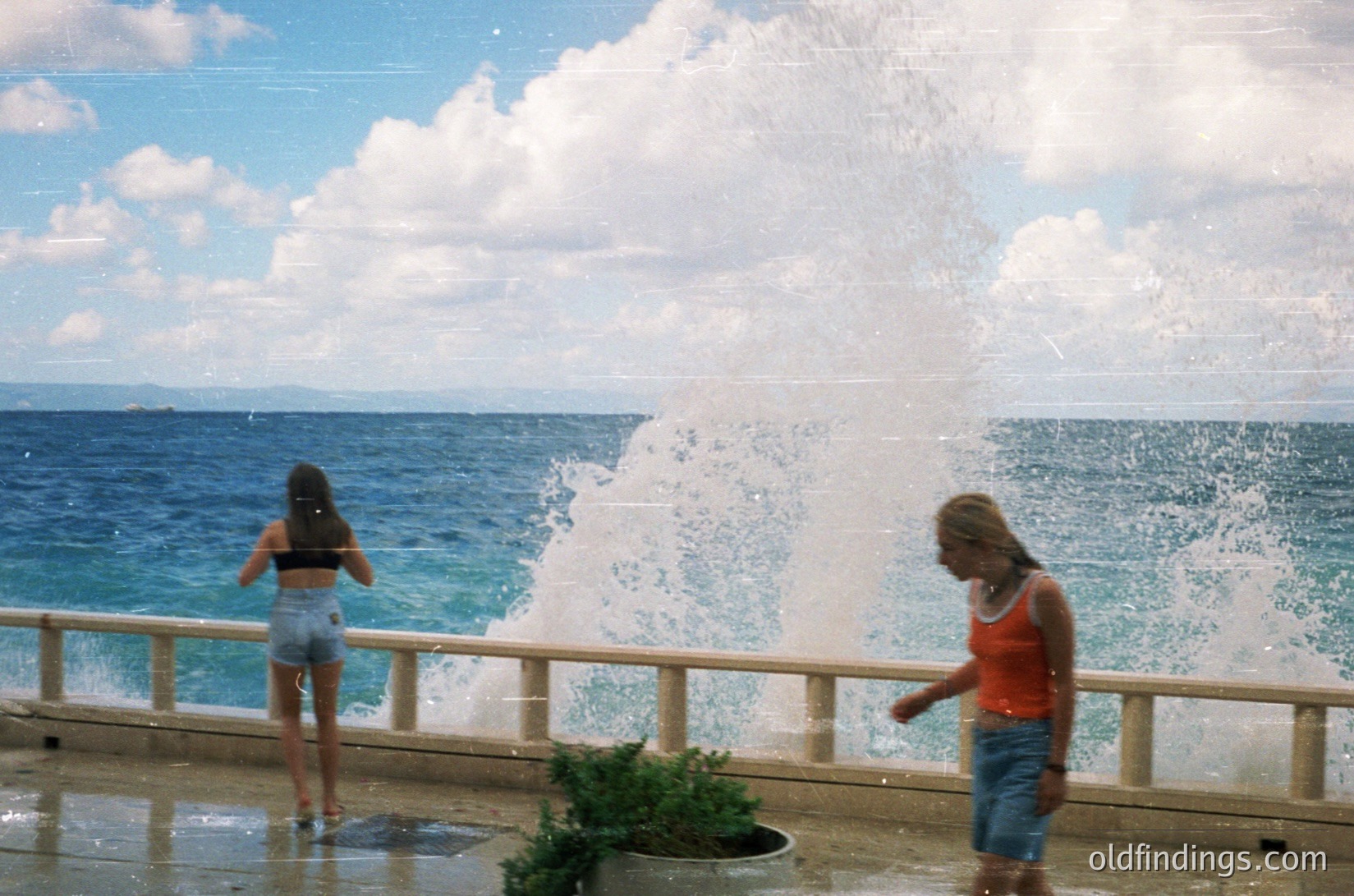 Two women enjoy a seaside fountain’s spray on a sunny day, likely mid-20th century. One wears a red tank top and denim shorts; the other, a bikini top and shorts. The ocean and wooden railing frame the scene, suggesting a resort or public promenade.