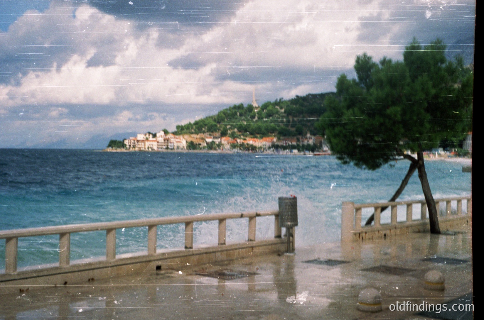 Vintage coastal scene featuring a seaside promenade with wooden railings and wet pavement. Distant hillside town with Mediterranean-style architecture and a lighthouse. Overcast sky with soft, diffused light. Likely Adriatic or Aegean region, mid-20th century.