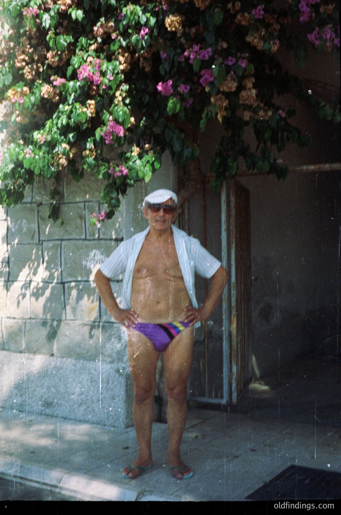 Vintage seaside portrait of an elderly man in mid-20th century swimwear—white shirt, purple shorts, and sunglasses—posed against a stone wall draped with flowering bougainvillea. Likely Mediterranean or coastal Europe, 1950s–1970s.