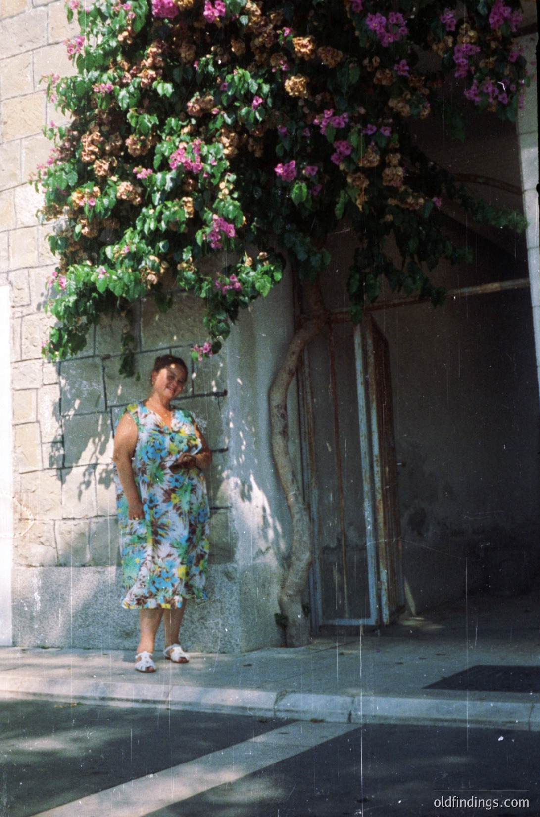 Vintage floral dress with geometric patterns stands out against blooming bougainvillea-covered wall. Concrete archway and weathered door suggest mid-20th-century European architecture. Soft focus hints at analog photography.