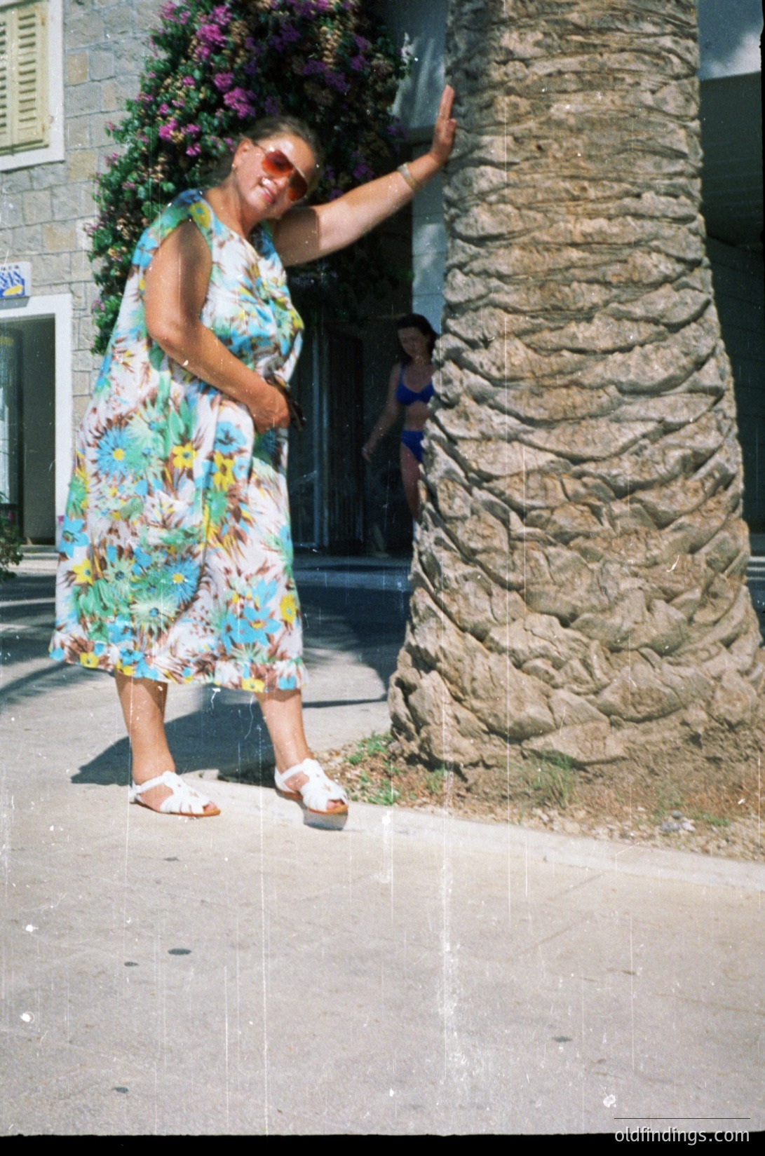 Vintage seaside portrait of woman in bold 1970s floral dress leaning against rustic stone pillar. Bright floral print and sunglasses suggest summer leisure. Background shows classic Mediterranean architecture with arched doorway and blooming vines.