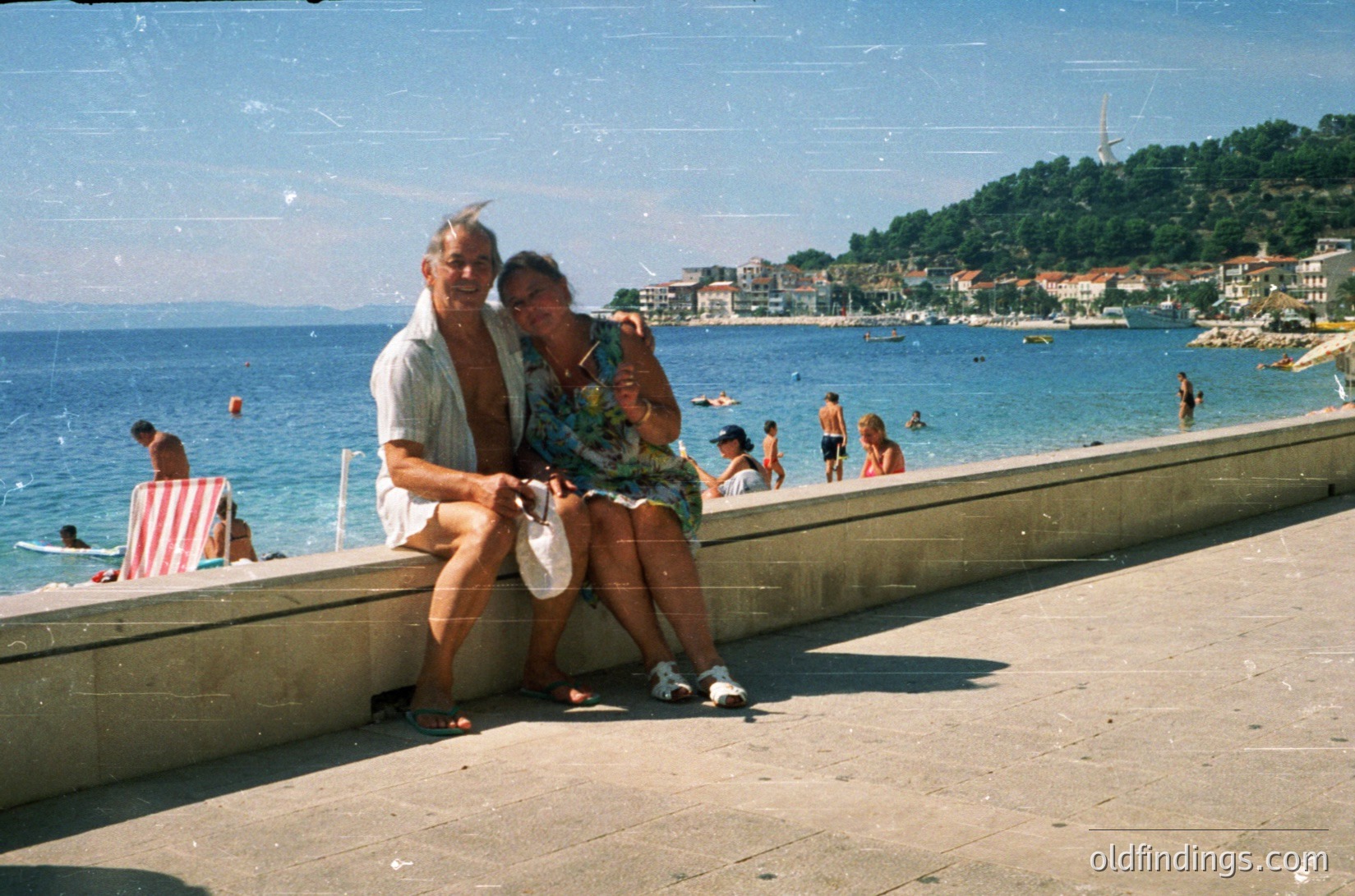 Couple posing on a concrete pier by a seaside resort, mid-20th century. Man in light linen shirt, woman in floral dress. Background shows coastal town with buildings, swimmers, and a hillside.