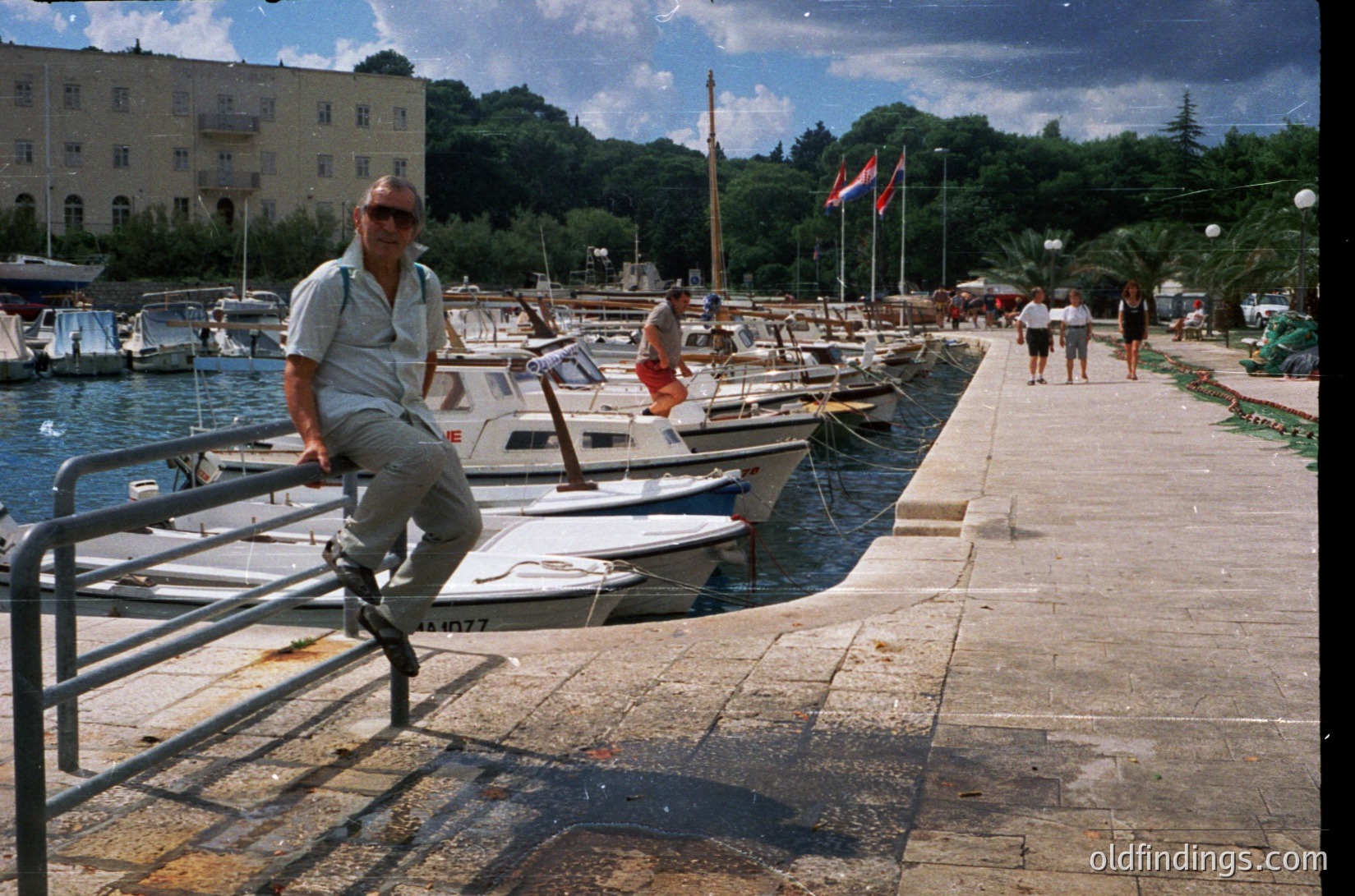 Man in retro sunglasses poses on a metal railing beside a marina with small boats docked in a seaside town. Soviet-era architecture and flags suggest Eastern European coastal vibes.
