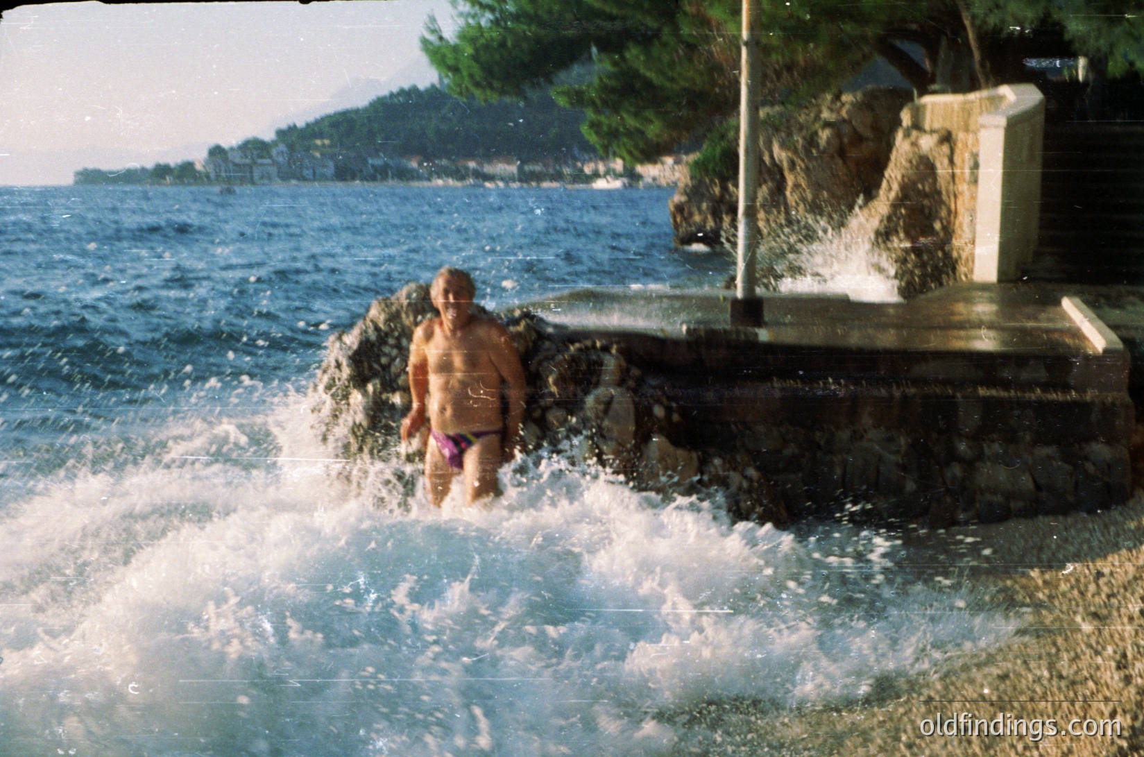 A person stands in shallow coastal waters near a concrete jetty, wearing a swimsuit with a floral pattern. The scene includes a rocky shoreline, waves crashing, and a distant hillside with scattered buildings. Likely Mediterranean seaside, 1970s–1990s era.