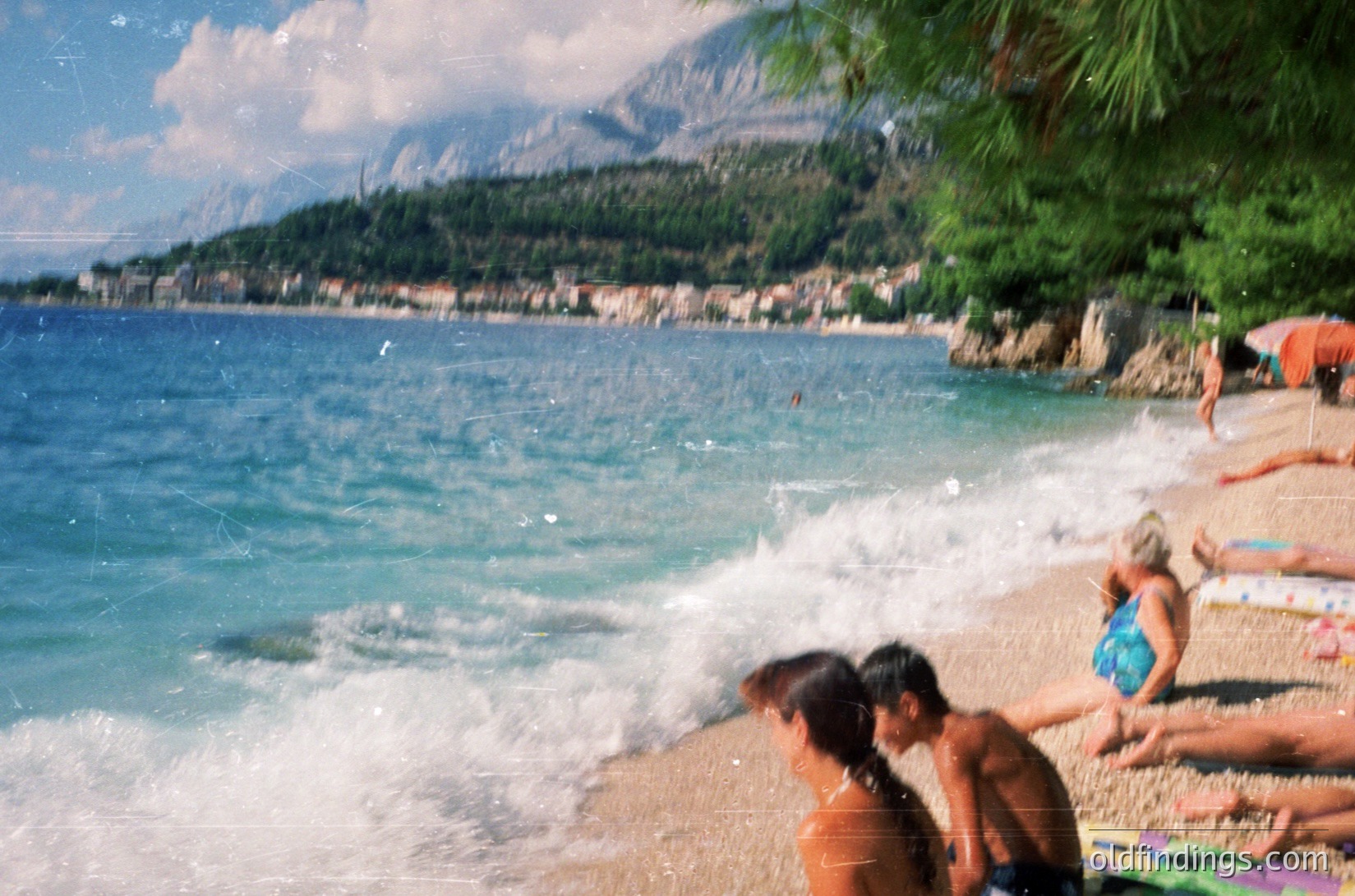 Coastal scene with pebbled beach and gentle waves crashing ashore. Mountainous backdrop with scattered buildings. Two people wading in shallow water; others sunbathing on towels. Lush greenery frames right edge. Likely Mediterranean or Adriatic region, summer season.