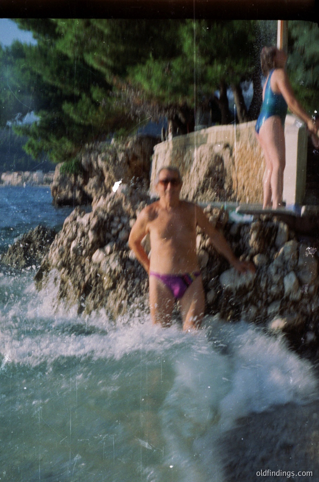 A man in purple swim trunks stands in a natural rock waterfall pool by a seaside cliff, with water cascading around him. Another person in a teal swimsuit stands on a concrete ledge behind. Lush greenery and rocky terrain frame the scene, suggesting a Mediterranean coastal setting.