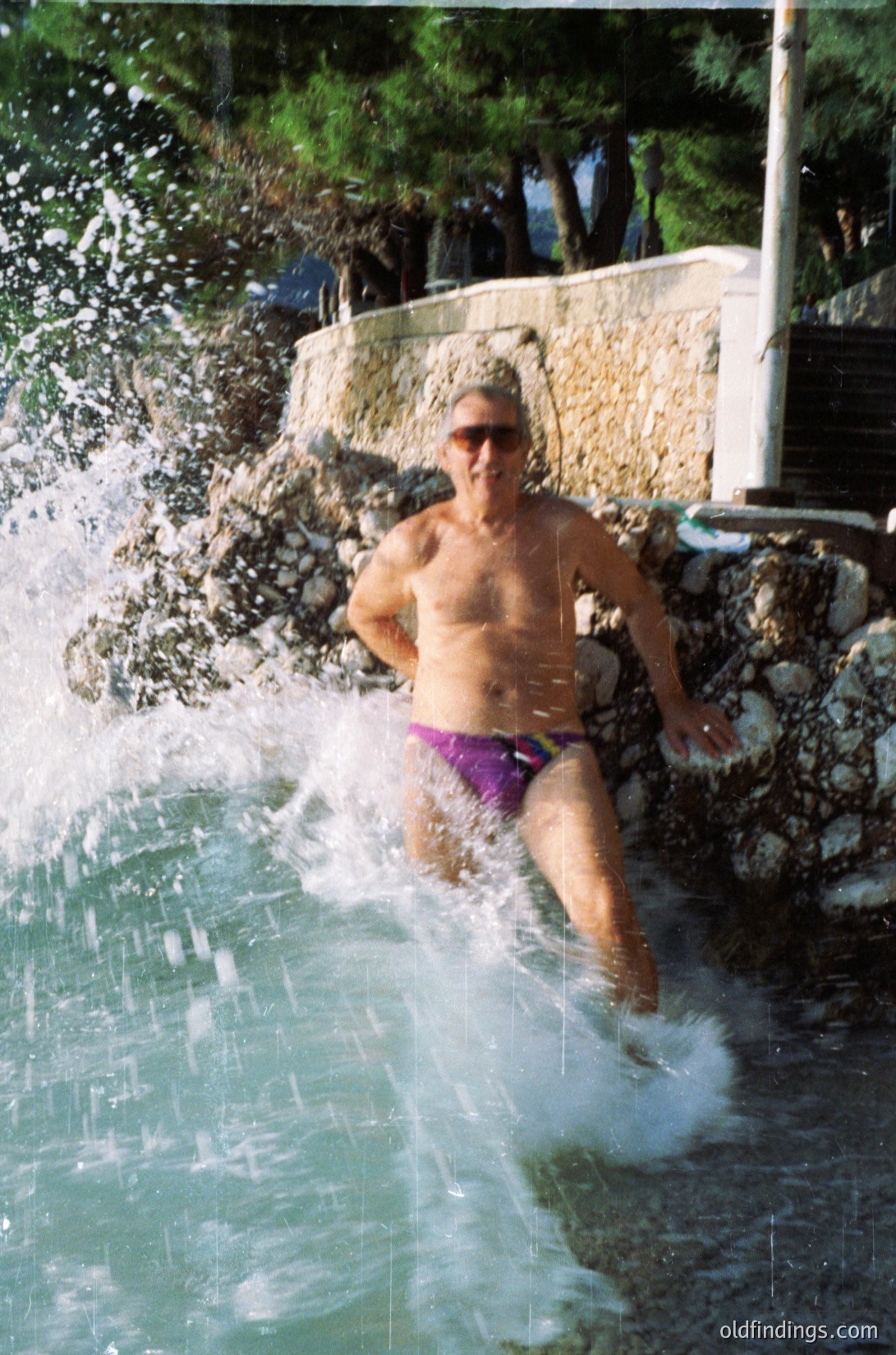 Man mid-dive into a cascading water feature, wearing purple swim trunks and sunglasses, set against a stone-lined pool edge. Lush greenery and stonework suggest a resort or garden setting. Likely late 20th century (1980s-1990s).