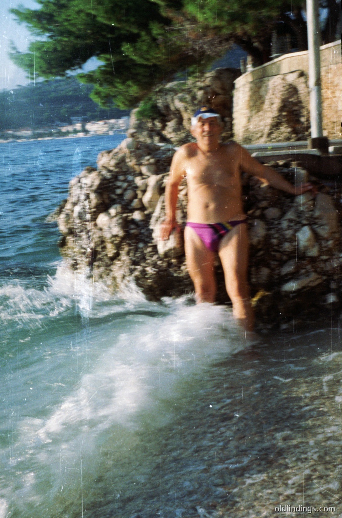 Man in bright pink swim trunks stands at rocky seaside ledge, mid-wave splash. Lush greenery and concrete steps visible in background. Likely Mediterranean coastal scene, 1970s-1990s era.