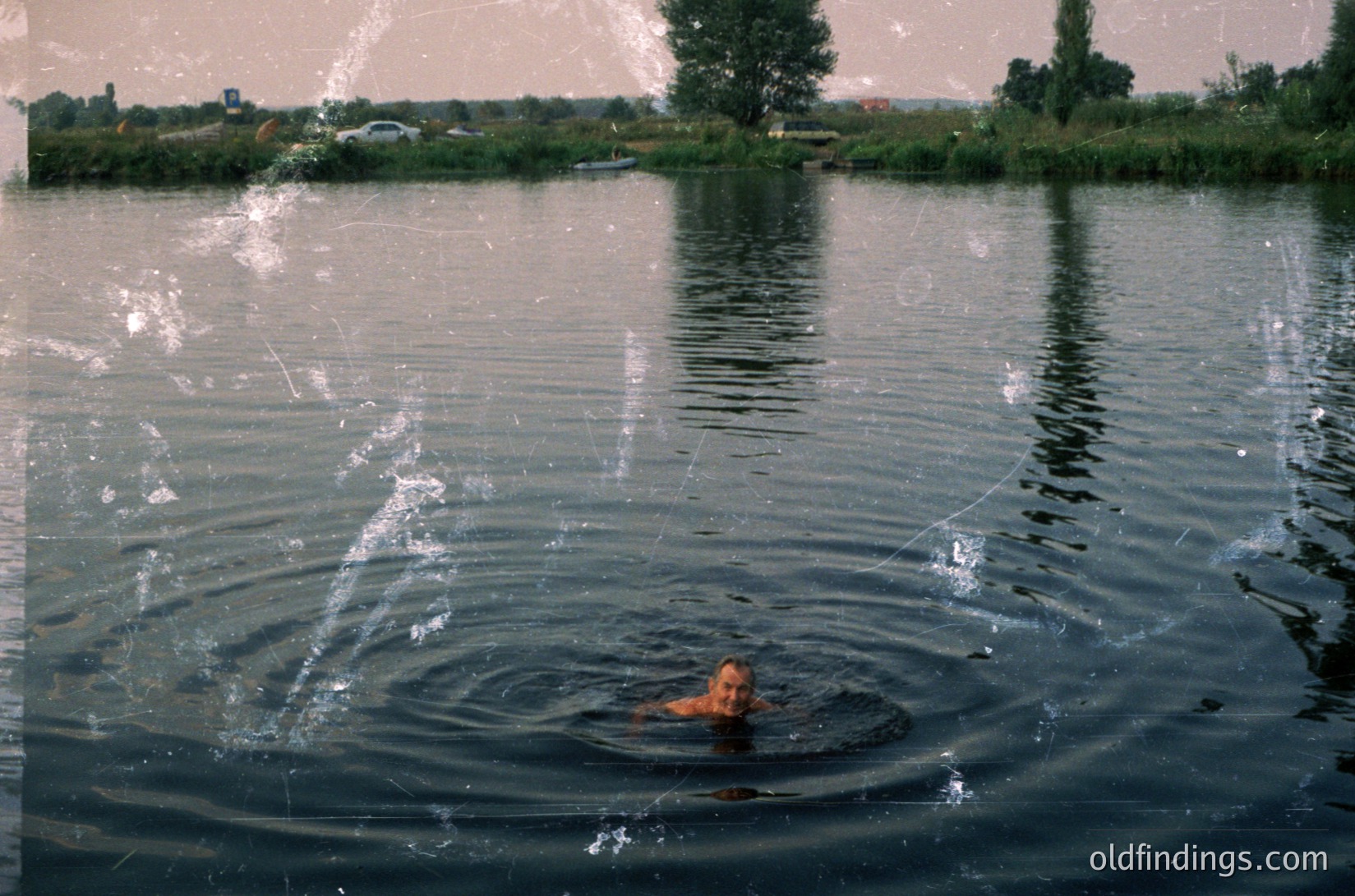 Vintage black-and-white photo of a lone figure swimming in calm, reflective water. Ripples spread outward from the swimmer’s head, creating a mirror-like surface. Distant shoreline shows sparse vegetation and a few structures, suggesting a rural or lakeside setting. Film grain and soft focus evoke mid-20th-century photography ().
