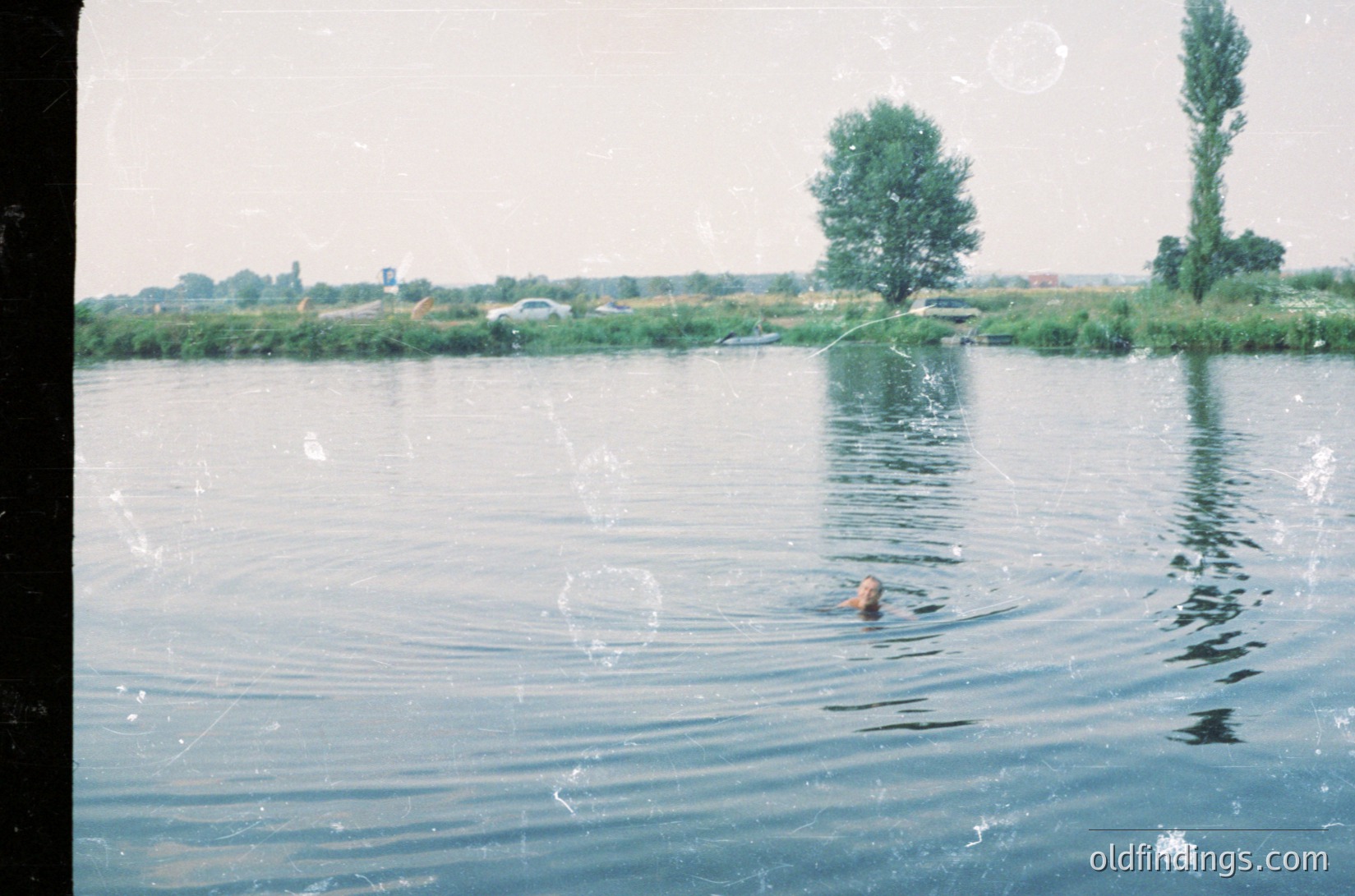 Vintage photo of a lone swimmer in a calm, reflective body of water with lush greenery and distant low-rise buildings. Likely mid-20th century due to film grain and clothing style.