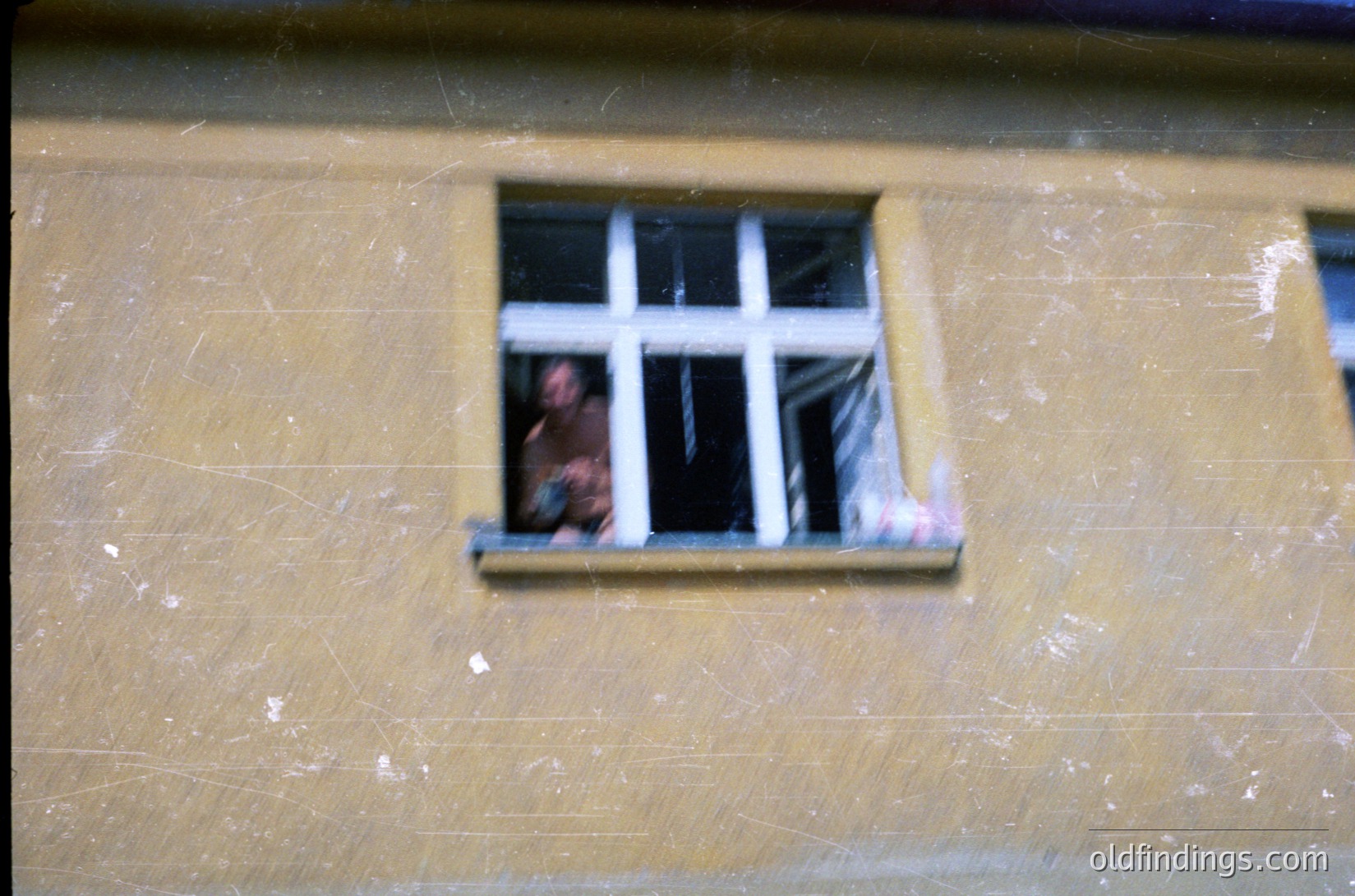 A close-up of a worn, yellowish plaster wall with a small, square window featuring six panes. Inside, a person’s face is partially visible, framed by the window bars. The scene suggests a confined or restricted space, possibly residential or institutional.
