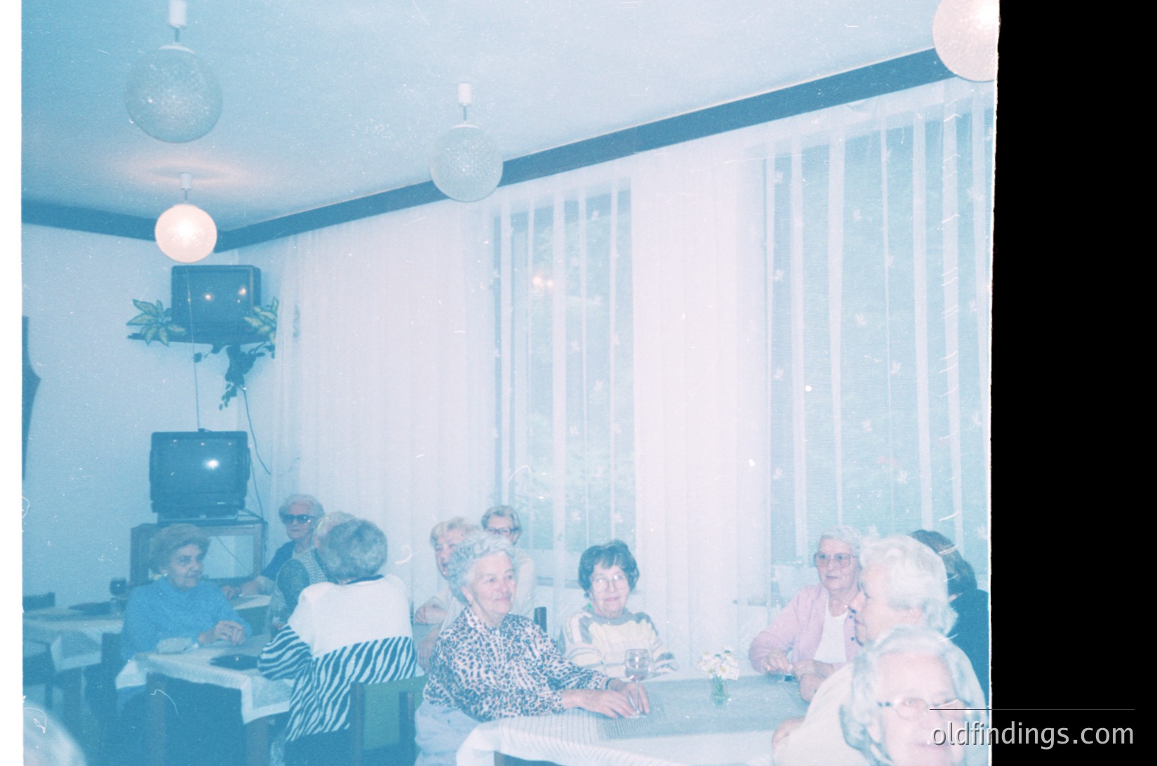 Vintage indoor gathering in a retro-style lounge, likely from the 1970s. Six elderly women seated around a table with floral arrangements, framed by sheer curtains and a small TV on a side table. Decor includes hanging plants and spherical pendant lamps. Warm, nostalgic atmosphere.