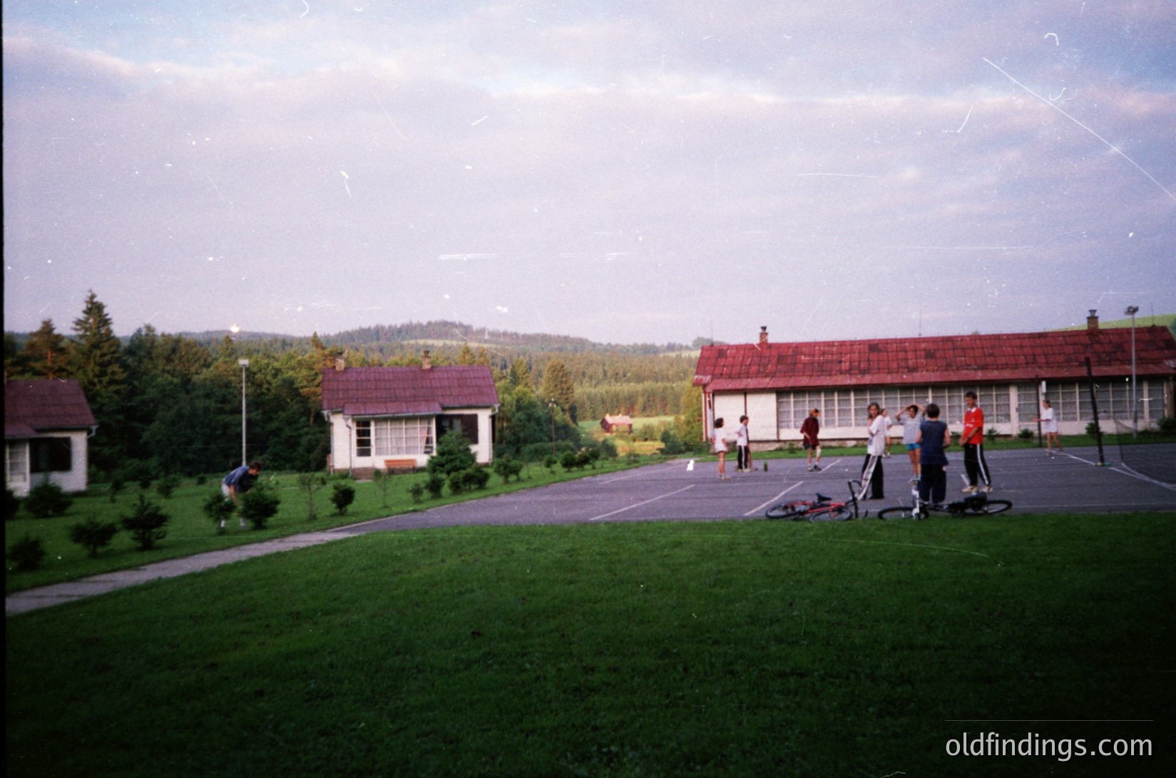 Mid-20th century residential complex with red-tiled roofs and white walls, set in a lush green landscape. Group of children playing on a paved area, some on bicycles. Low-lying hills and trees in background. Likely Eastern European suburban setting, 1960s-1980s.