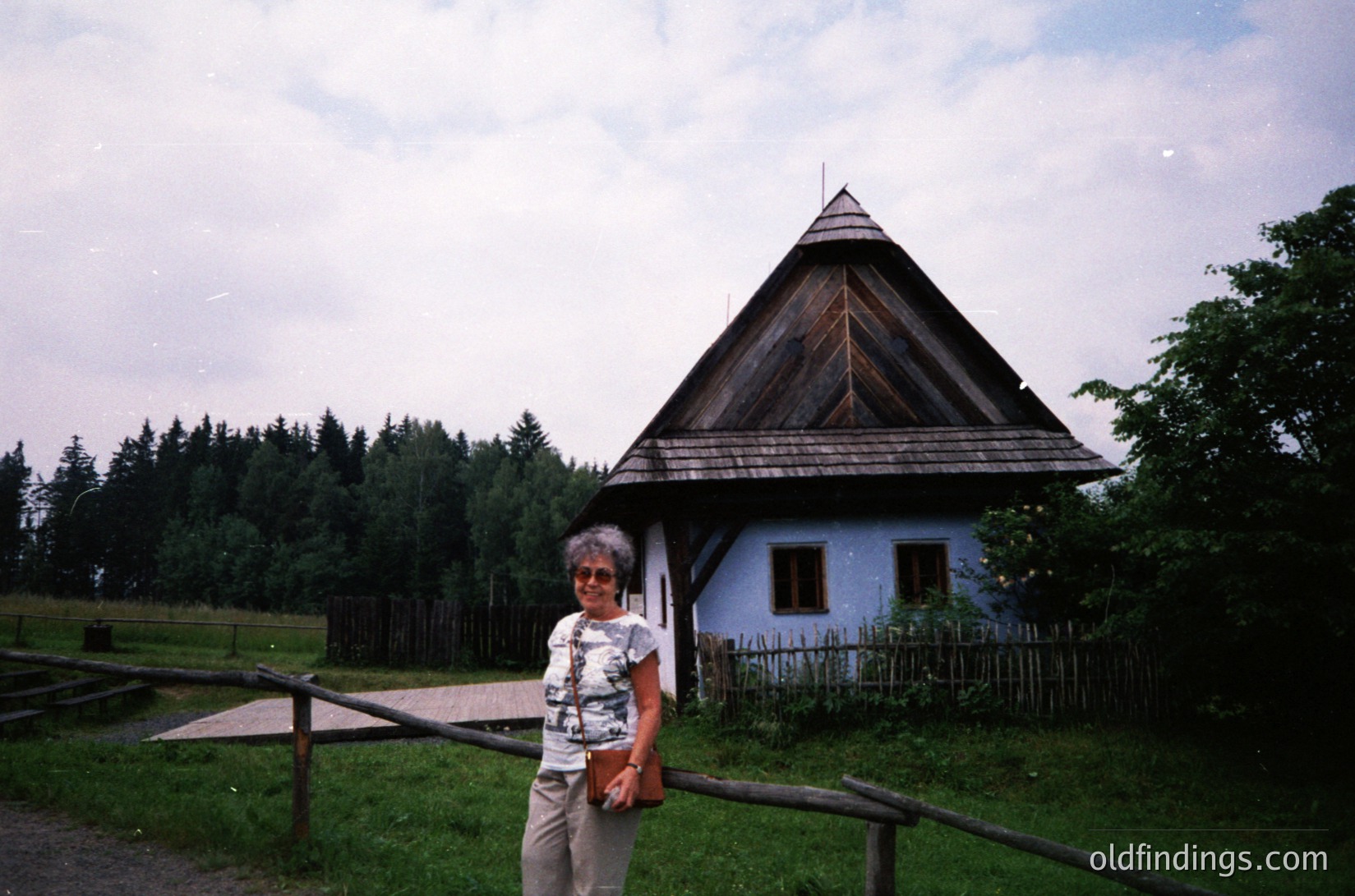 Traditional timber-framed cottage with steep gabled roof, set in a rural forest clearing. Woman in patterned blouse and beige pants poses near wooden fence. Likely Eastern European folk architecture, mid-20th century.