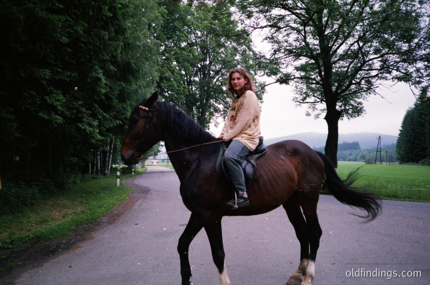 Woman riding a dark brown horse along a tree-lined road, dressed in a beige jacket and jeans. Lush greenery and misty hills in background suggest a rural, possibly European countryside setting. Image likely captured in the late 20th century.