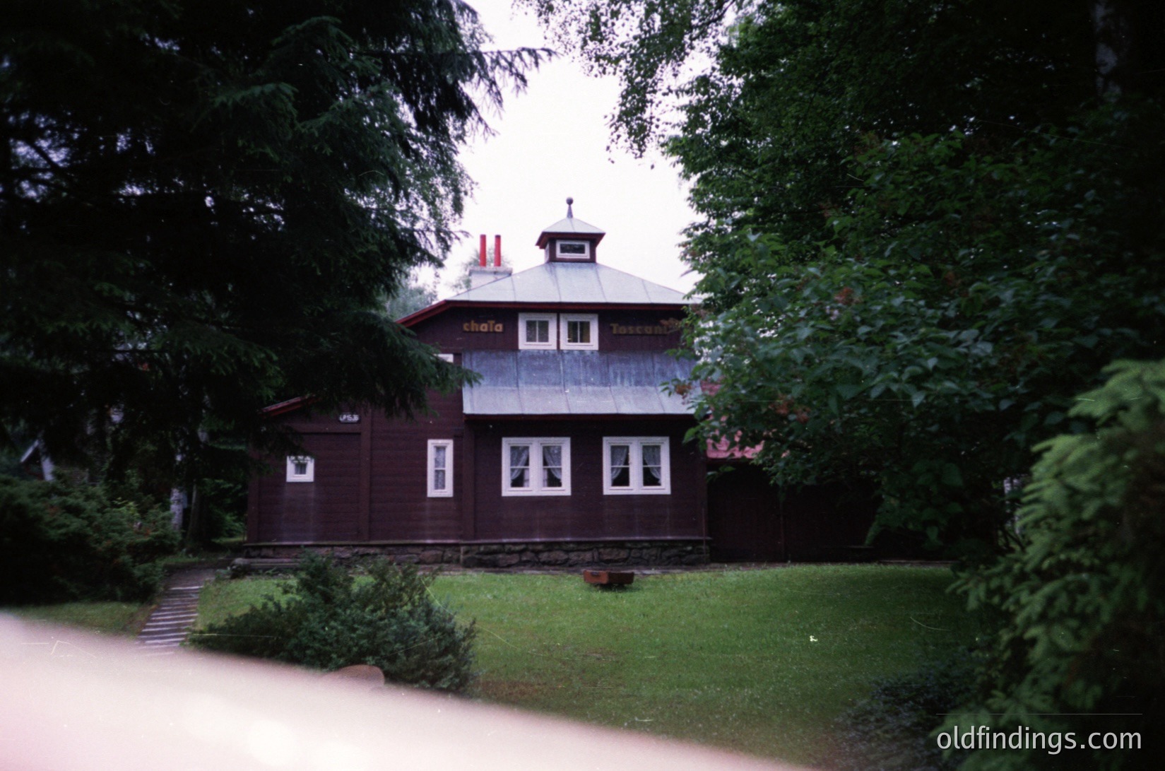 Two-story wooden building with a gabled roof, featuring a small tower-like extension on the upper floor. The signage "Chalet" and "Tanev" suggests a resort or lodge. Surrounded by dense greenery and a paved pathway. Likely Eastern European alpine or forest setting.
