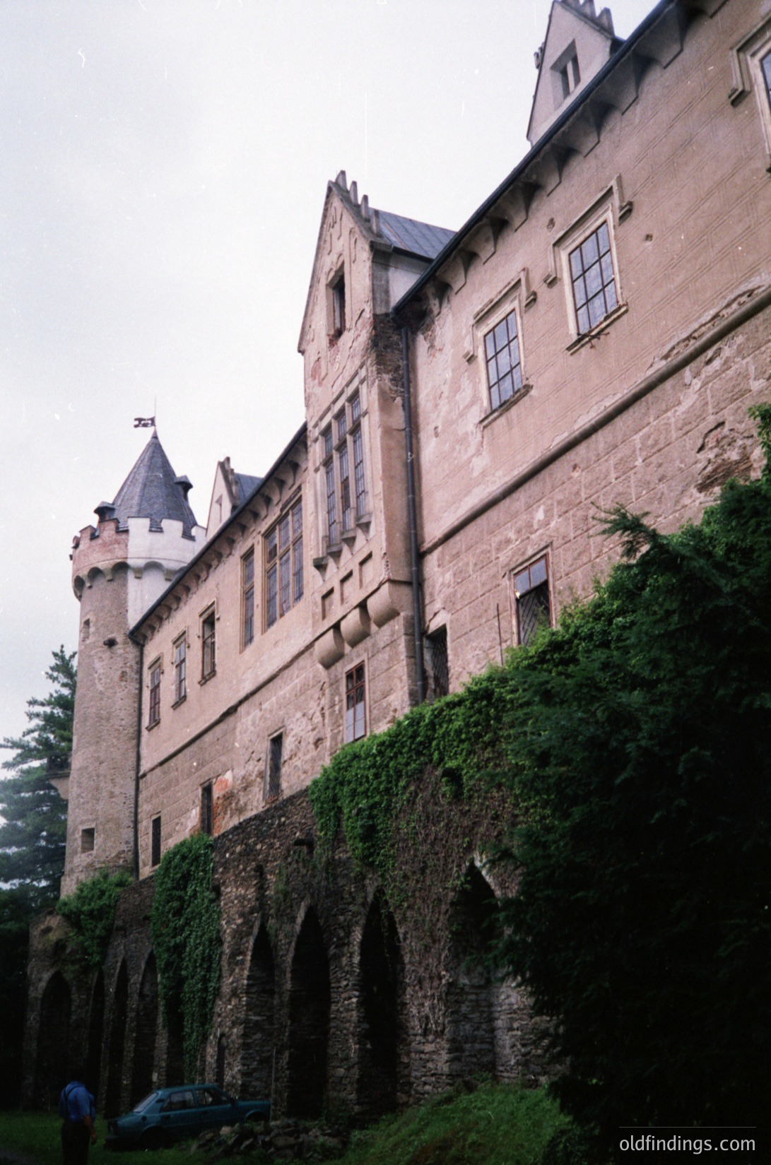 Gothic Revival castle with crenellated towers, ivy-covered stone walls, and arched buttresses. Likely European, 19th-century architectural style. Overcast sky enhances dramatic texture.