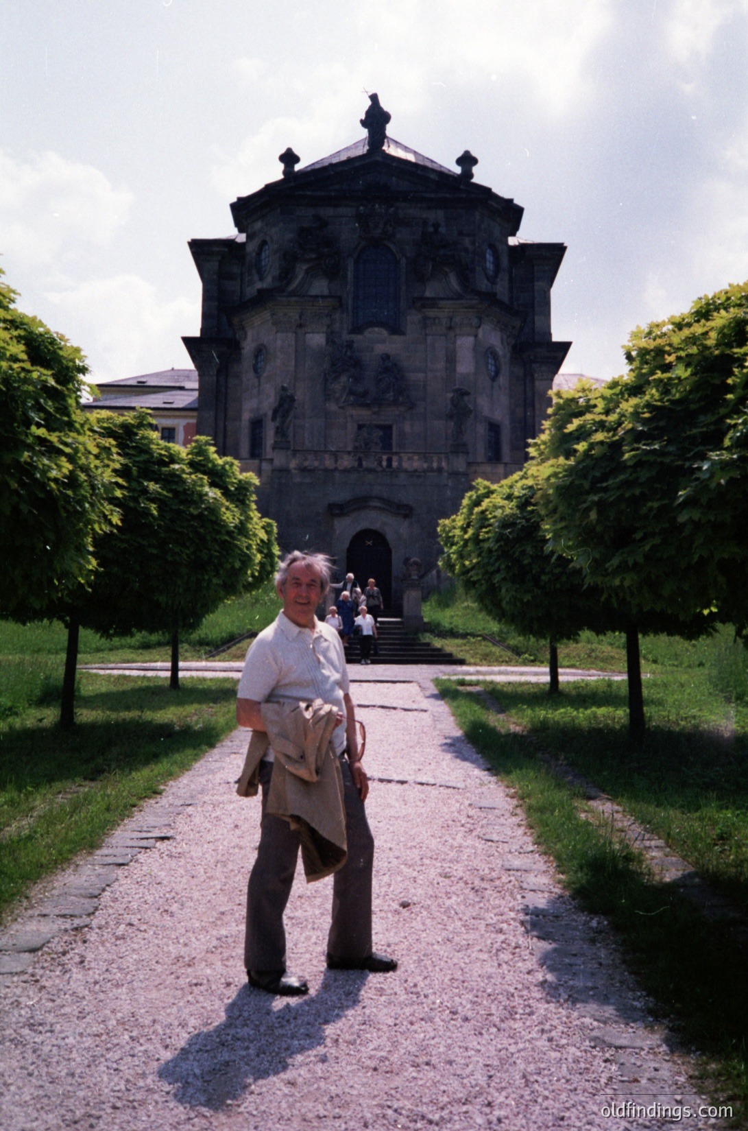 Neoclassical stone gatehouse with sculpted reliefs and a triangular pediment, flanked by symmetrical tree-lined pathways. Man in 1970s attire (beige jacket, white shirt) stands mid-path, holding a bag. Lush greenery and cobblestone road enhance historic European park setting.