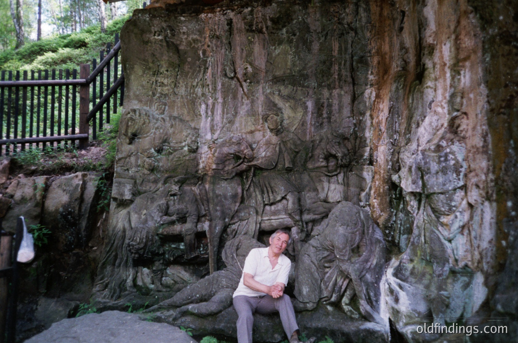 Monolithic stone relief sculpture depicting reclining figures in a natural rock setting, likely a Soviet-era public art installation. The man in mid-20th-century attire poses beside the artwork, suggesting a cultural or historical site.