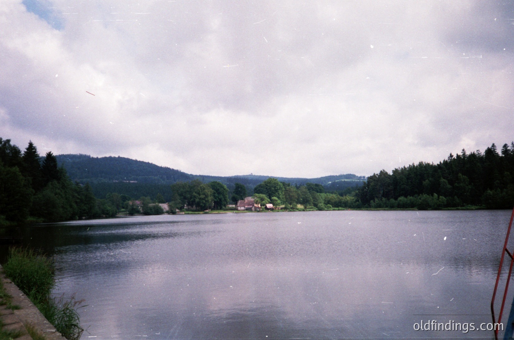 Lake scene with forested hills in background, calm water reflecting muted light. Rustic wooden structures along shoreline suggest rural European setting. Overcast sky enhances tranquil, timeless atmosphere. Likely 1980s-1990s analog photography.