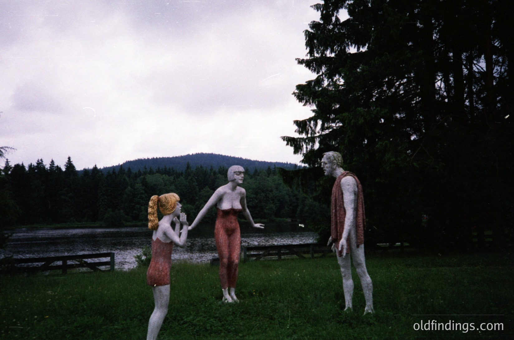 Vintage sculpture installation featuring three human-like figures in outdoor setting. Left figure wears a red dress, center figure holds hands with a blonde figure in red shorts. Natural lake and forested hills in background. Likely 1960s–1970s art piece.
