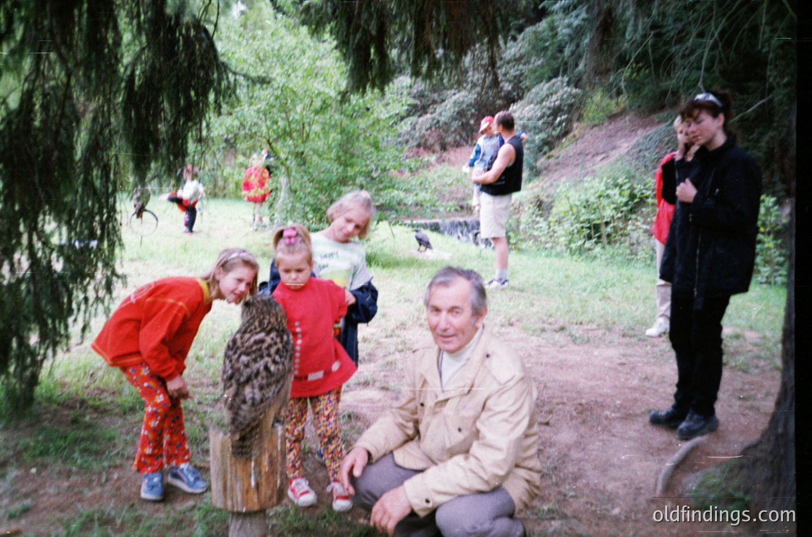 Vintage outdoor group photo in a wooded area, featuring a man in a beige jacket squatting with two young girls in patterned clothing. Others stand in the background, some holding cameras. Soft focus and natural lighting suggest a family or community gathering, likely mid-20th century.