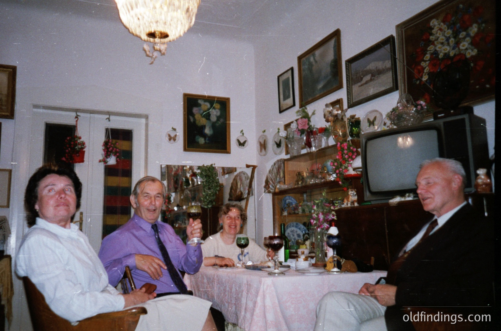Four adults gather in a mid-century living/dining space, adorned with floral wallpaper, vintage chandelier, and framed artwork. A small TV sits atop a shelved sideboard decorated with ceramic figurines. Glassware and a wine bottle suggest a social gathering, likely 1970s–1980s.