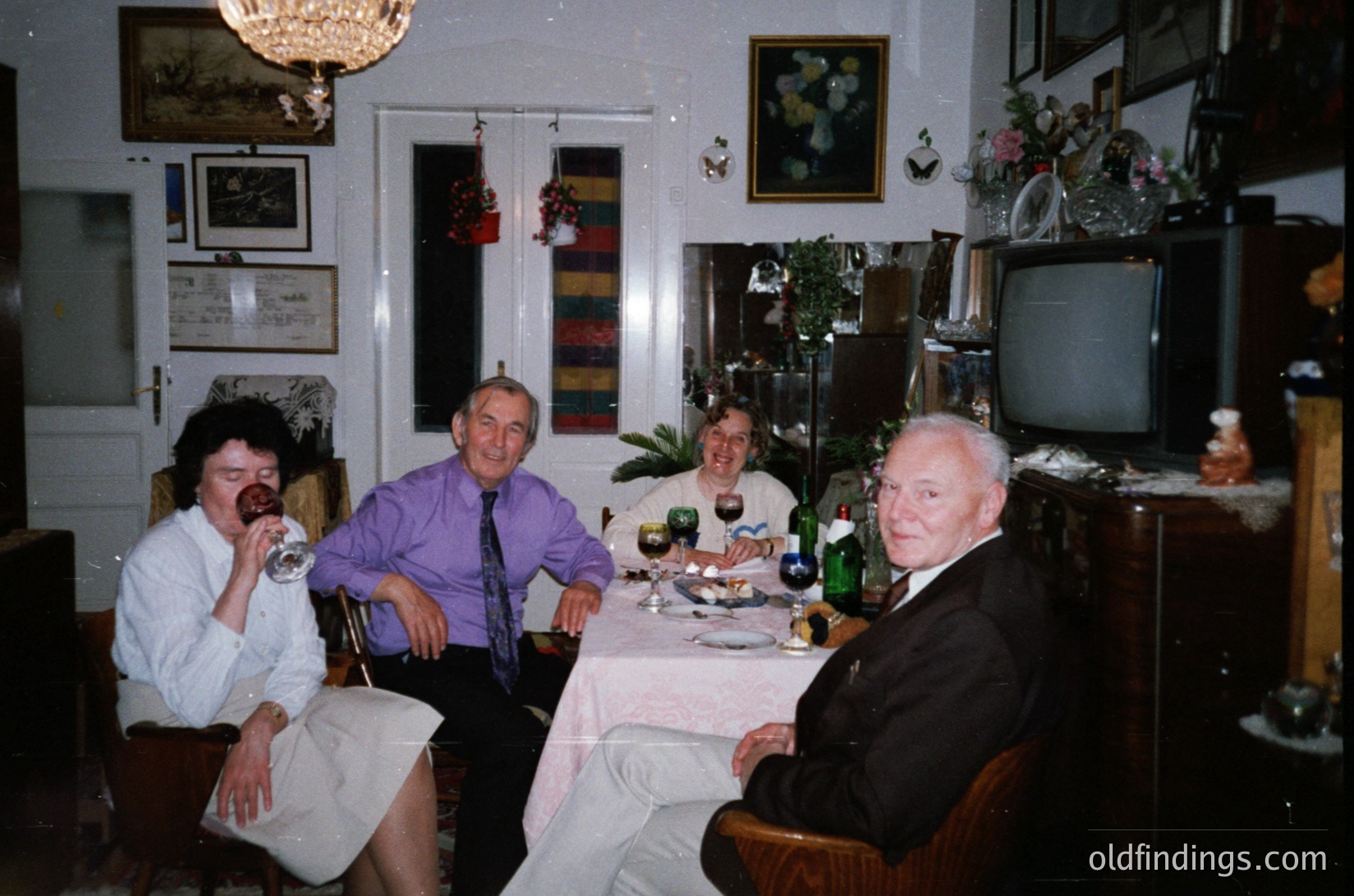 Vintage indoor gathering in a mid-century living/dining space. Four adults seated around a table with wine glasses, bottles, and a floral centerpiece. Decor includes framed art, a vintage TV, and a striped wall hanging. Formal attire suggests a special occasion, likely late 20th century (1970s–1990s).