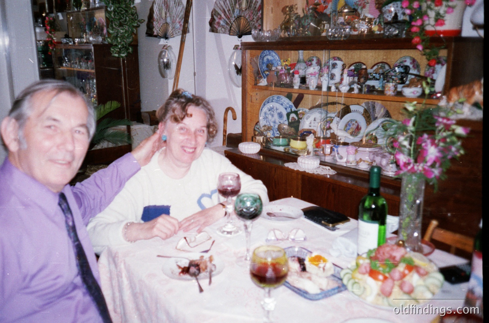 Vintage indoor dining scene featuring two adults seated at a table adorned with a spread of meats, cheeses, and wine. Wooden cabinets display blue-and-white ceramic plates. The man wears a purple shirt; the woman, a white blouse with a blue accessory. Floral arrangements and a potted plant add decor. Likely 1980s-1990s European home setting.