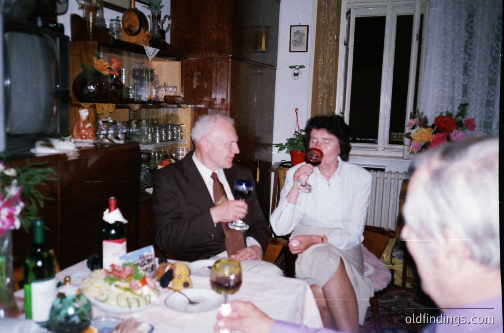 Vintage indoor gathering in a mid-century home, featuring two adults seated at a table adorned with wine bottles, glasses, and a decorated cake. The man wears a suit, while the woman in a white blouse holds a wineglass. Wooden cabinets and floral arrangements add to the 1960s–1970s ambiance.